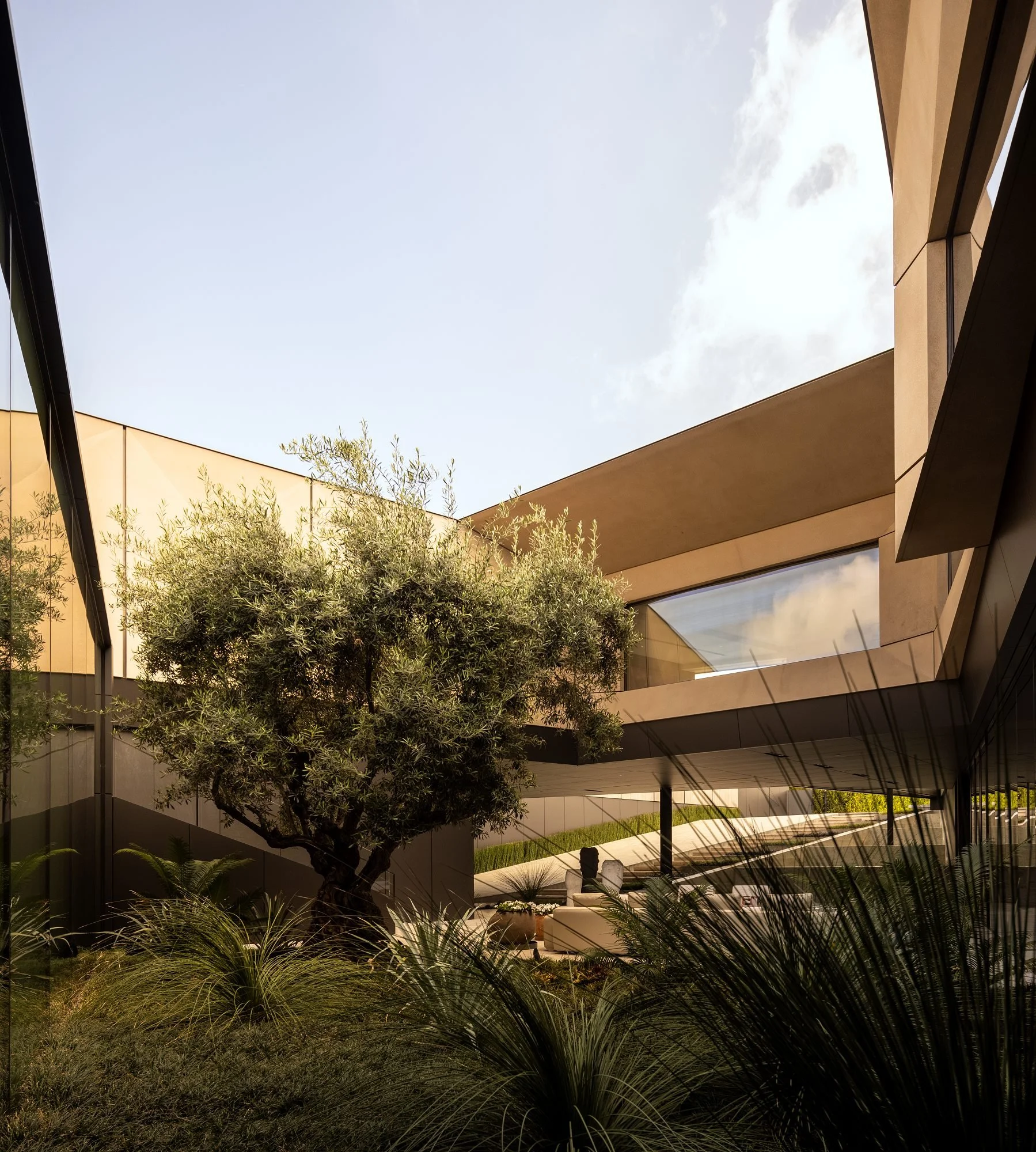 Modern building with large glass windows and beige exterior, trees, and landscaping in the foreground, clear sky with clouds.