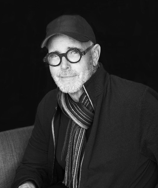 A grayscale portrait of a man with glasses, a baseball cap, and a striped scarf, sitting against a dark background.