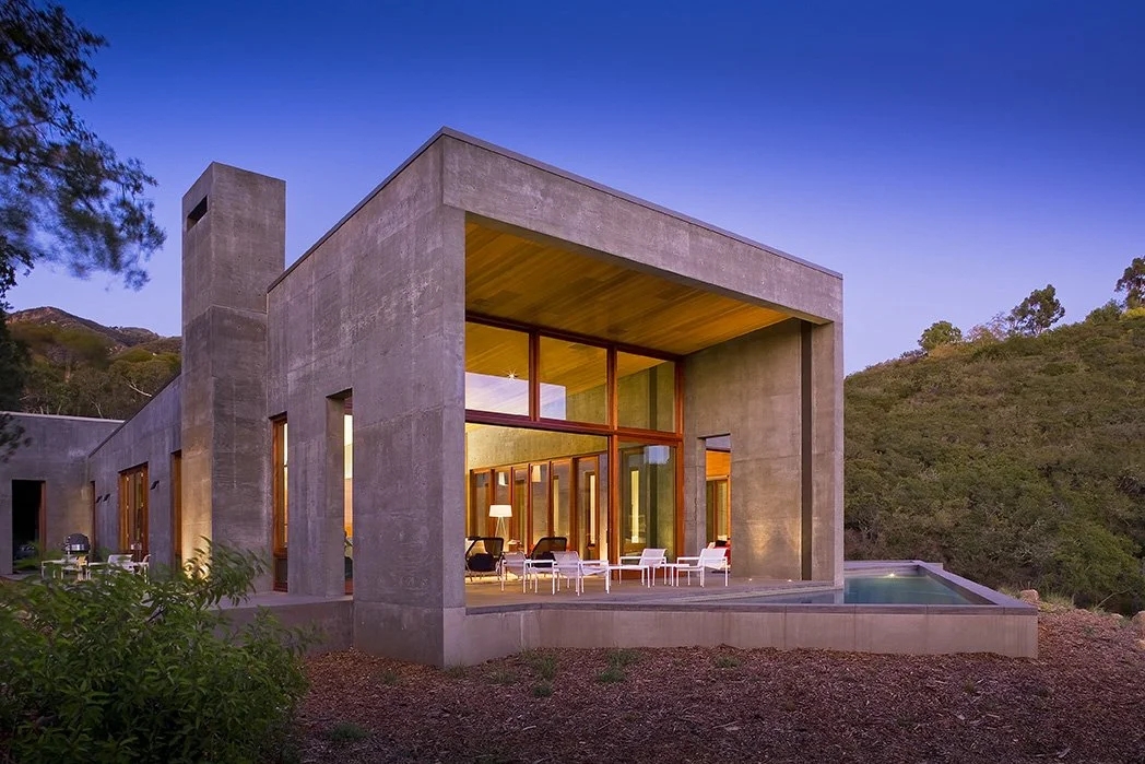 Modern concrete house with large windows and a patio, situated on a hillside with trees and a mountain in the background, during dusk.