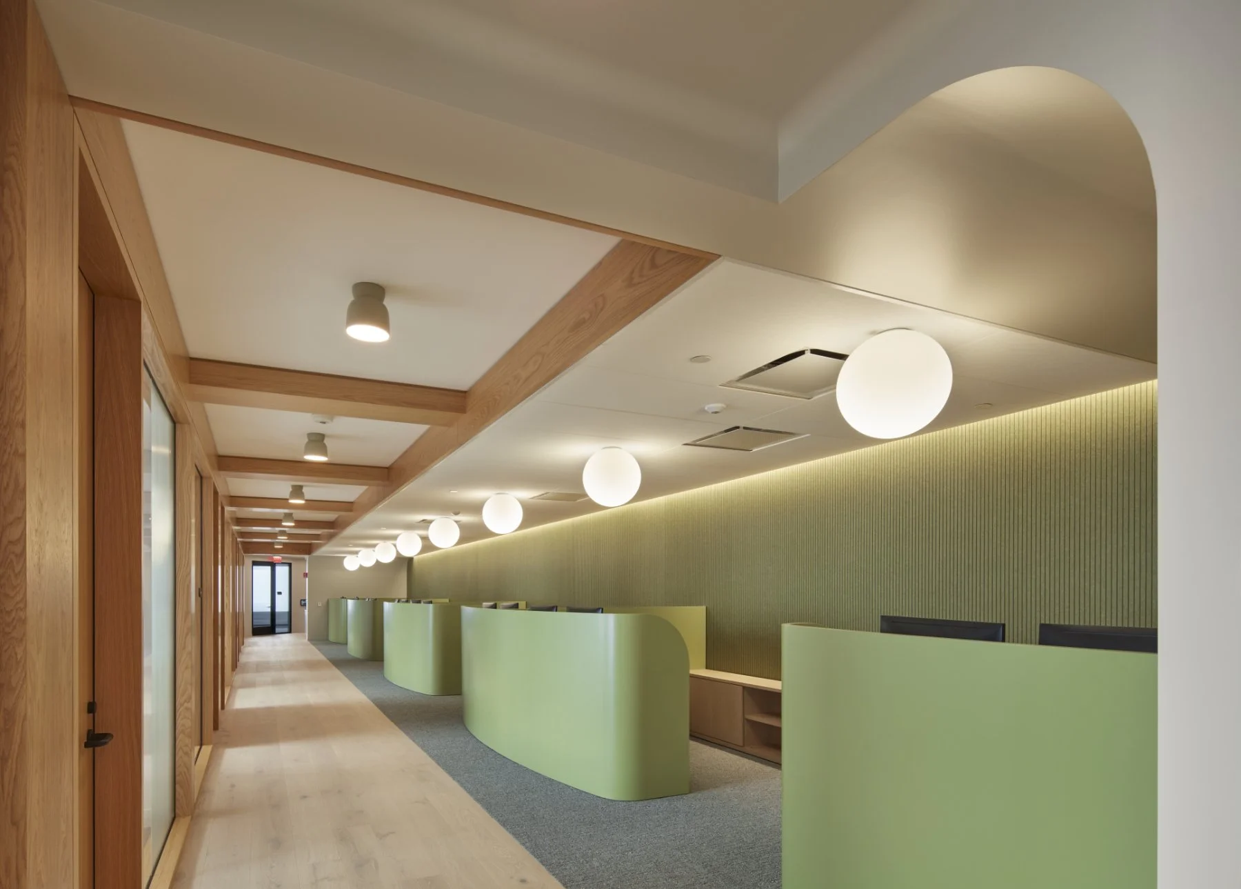 Interior of a modern office or lobby area with green curved partition walls, a wood-paneled ceiling, and spherical overhead lights, with a door at the end of the corridor.