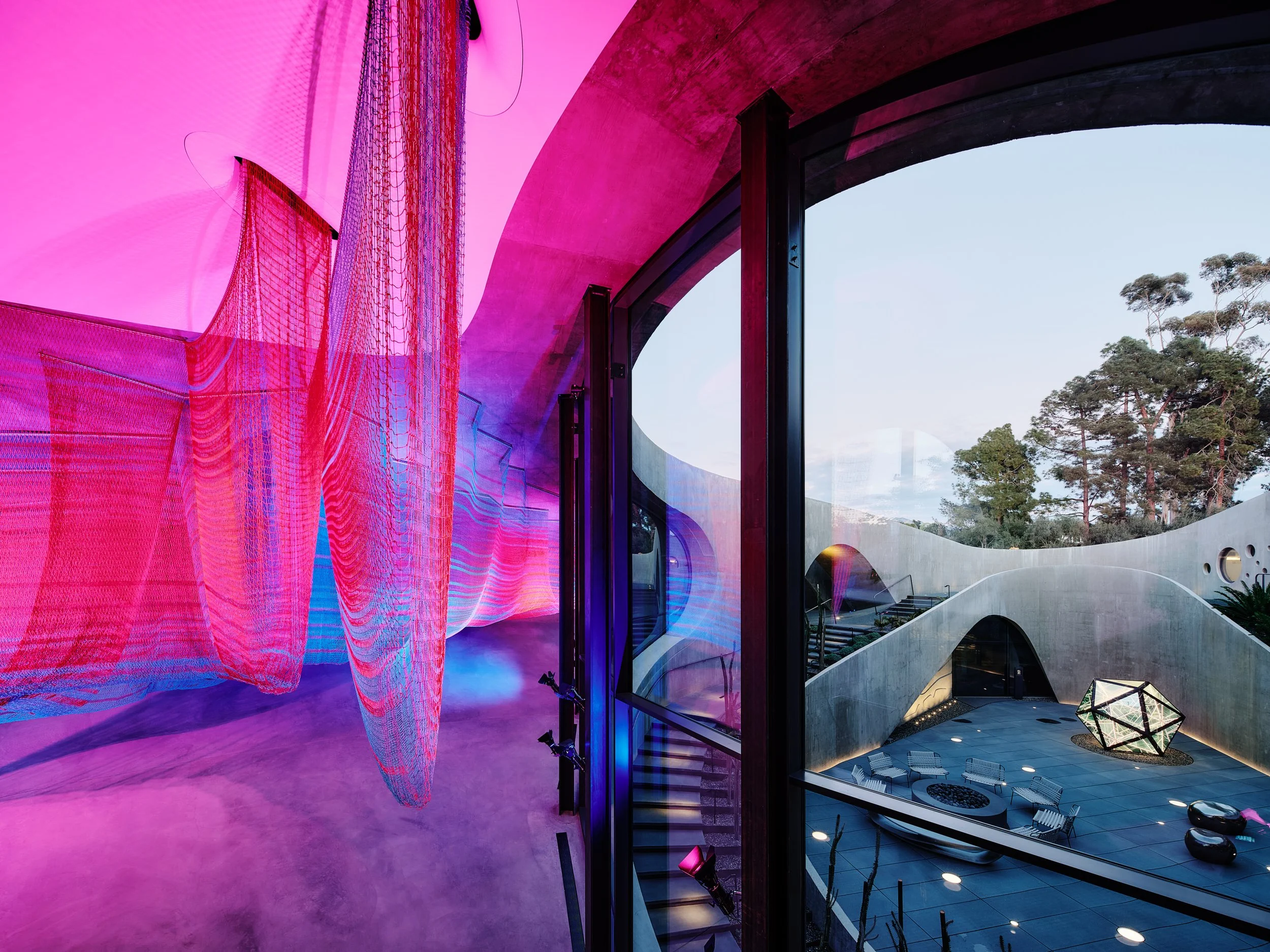Interior view of a modern building with vibrant pink and purple lighting, curved architecture, floor-to-ceiling windows, outdoor trees, and contemporary furniture.