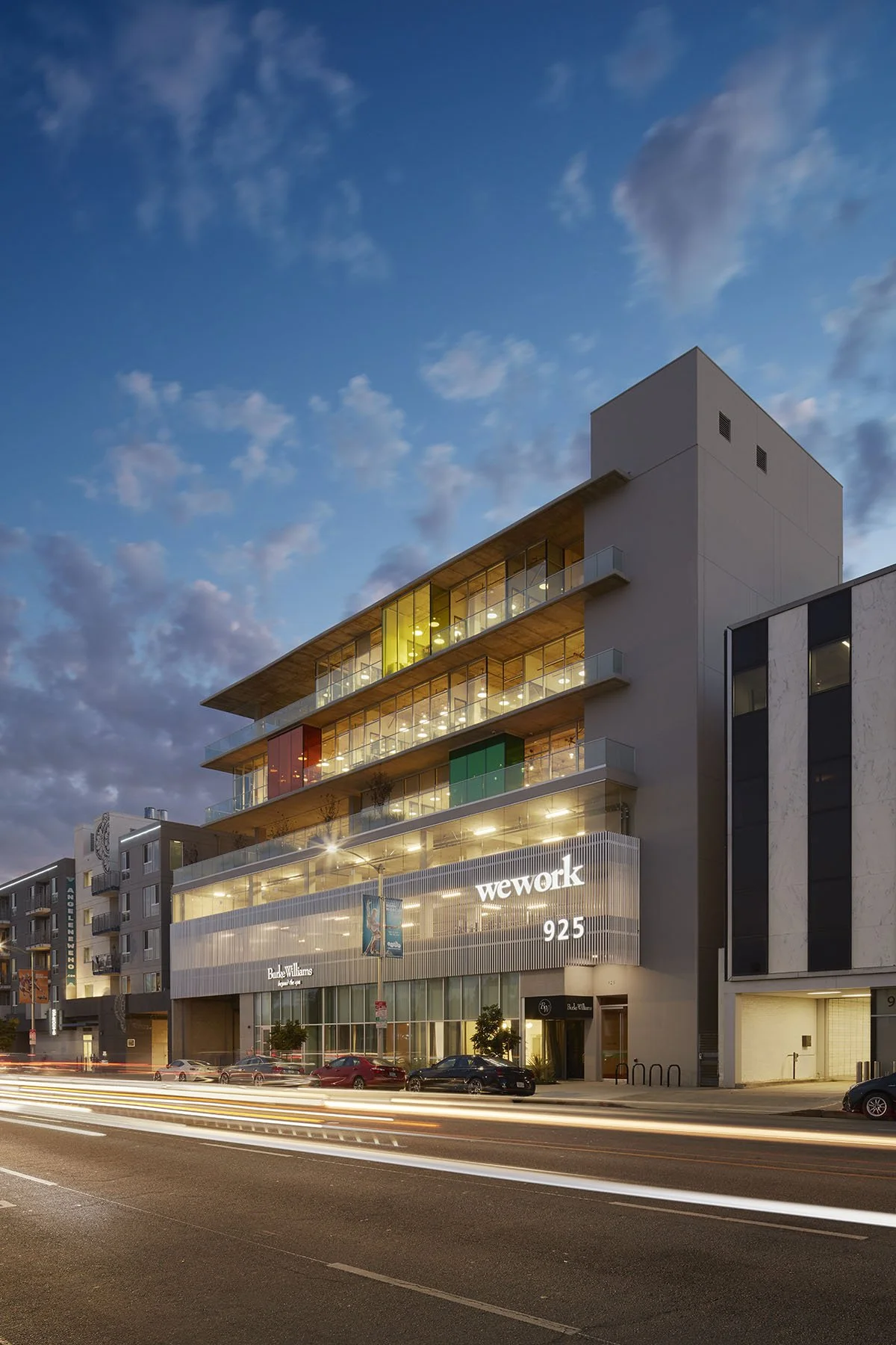 Modern multi-story office building with glass balconies at night, illuminated signage for 'WeWork' and 'Beke Williams,' and a busy street with car light trails in front.