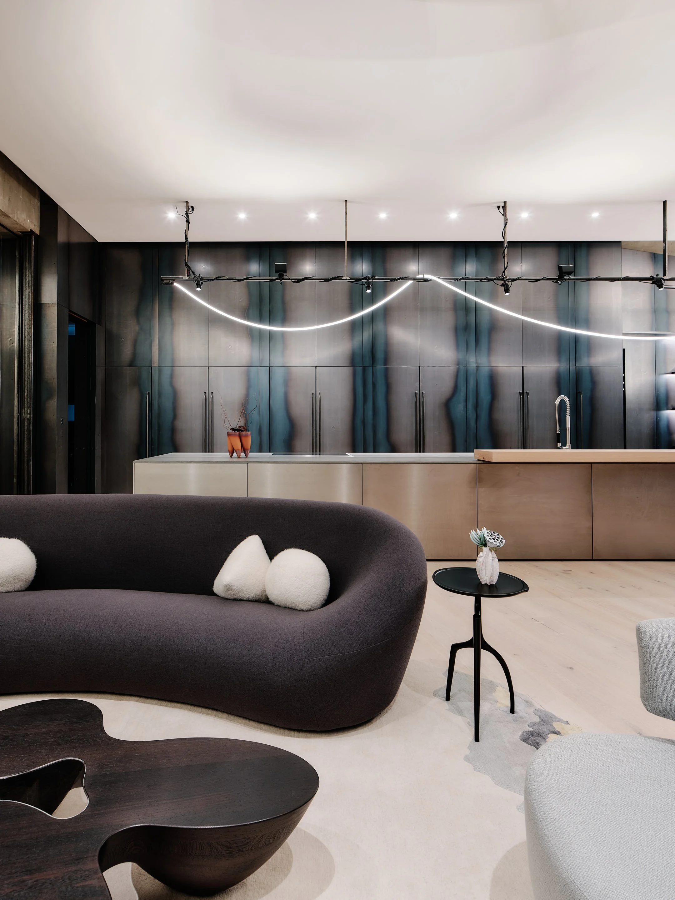 Modern living room with a curved gray sofa, white pillows, a black side table with a vase, a light-colored floor, and a dark textured wall with a light fixture design.