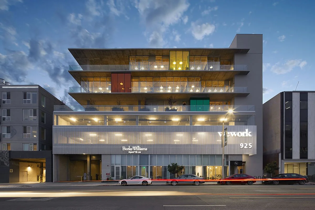 Modern multi-story building with glass balconies, illuminated interior, and signage for Burke Williams and Vewok at 925 address, with cars parked in front and a blue sky background at dusk.