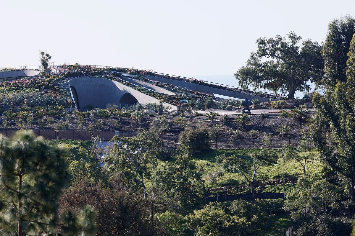 View of a hillside with a modern, curved building and surrounding landscaped greenery, including trees and plants.