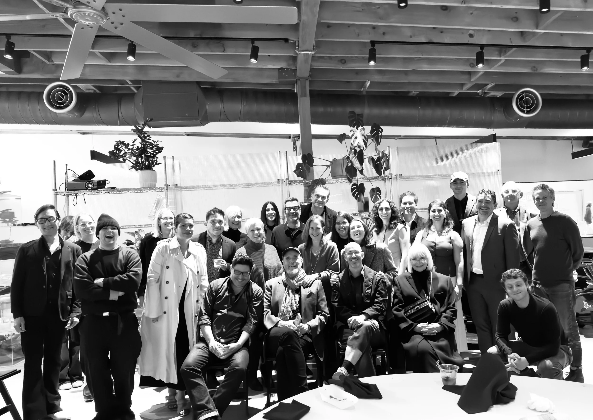 A large group of people gathered indoors, posing for a photo. The background shows an industrial-style ceiling with track lighting, a ceiling fan, and large ductwork. There are plants and shelves with office items behind the group.