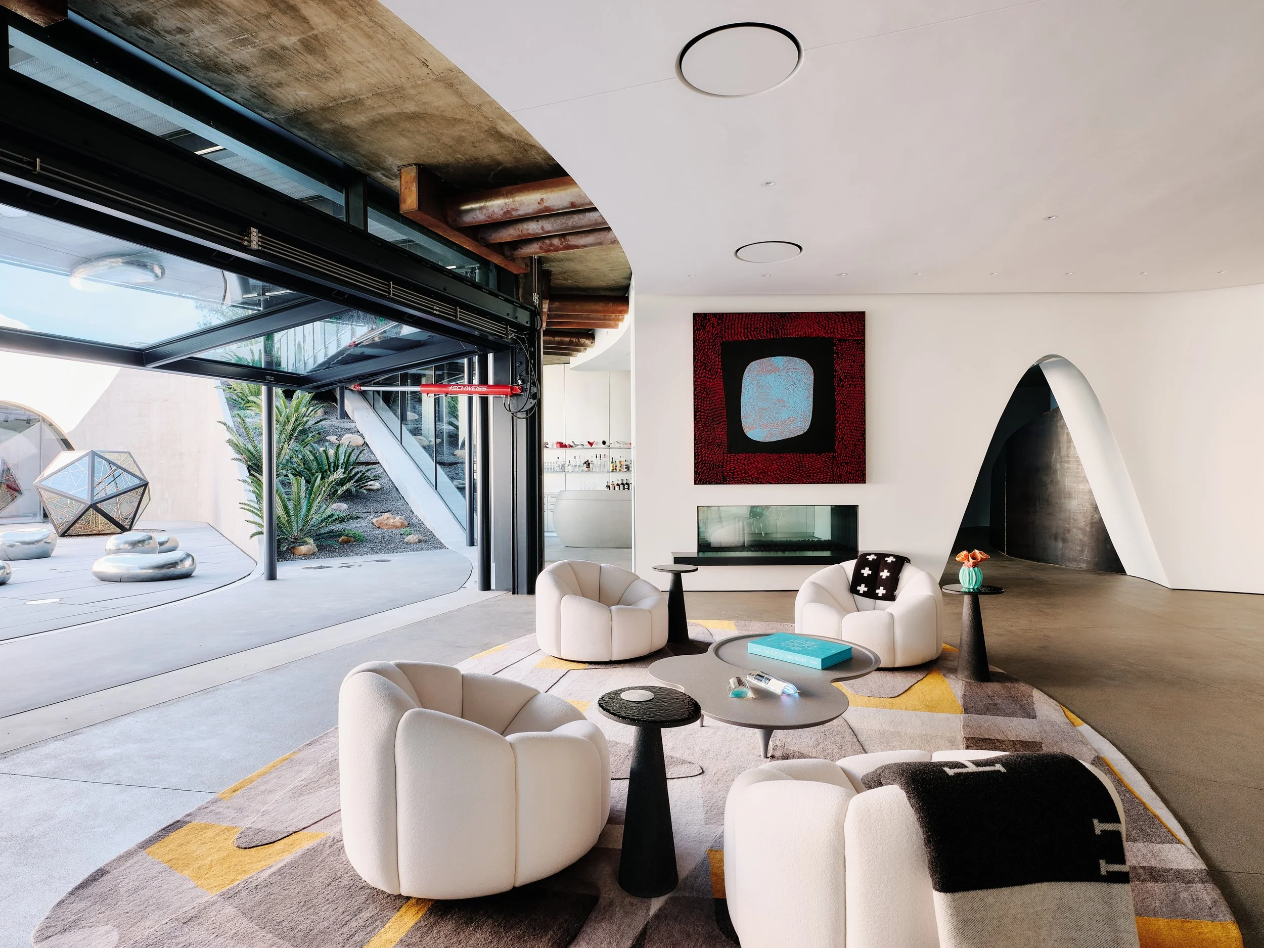 Modern living room with white rounded armchairs, black side tables, colorful area rug, and a large abstract artwork on the wall. Large windows show an outdoor space with plants and rocks.