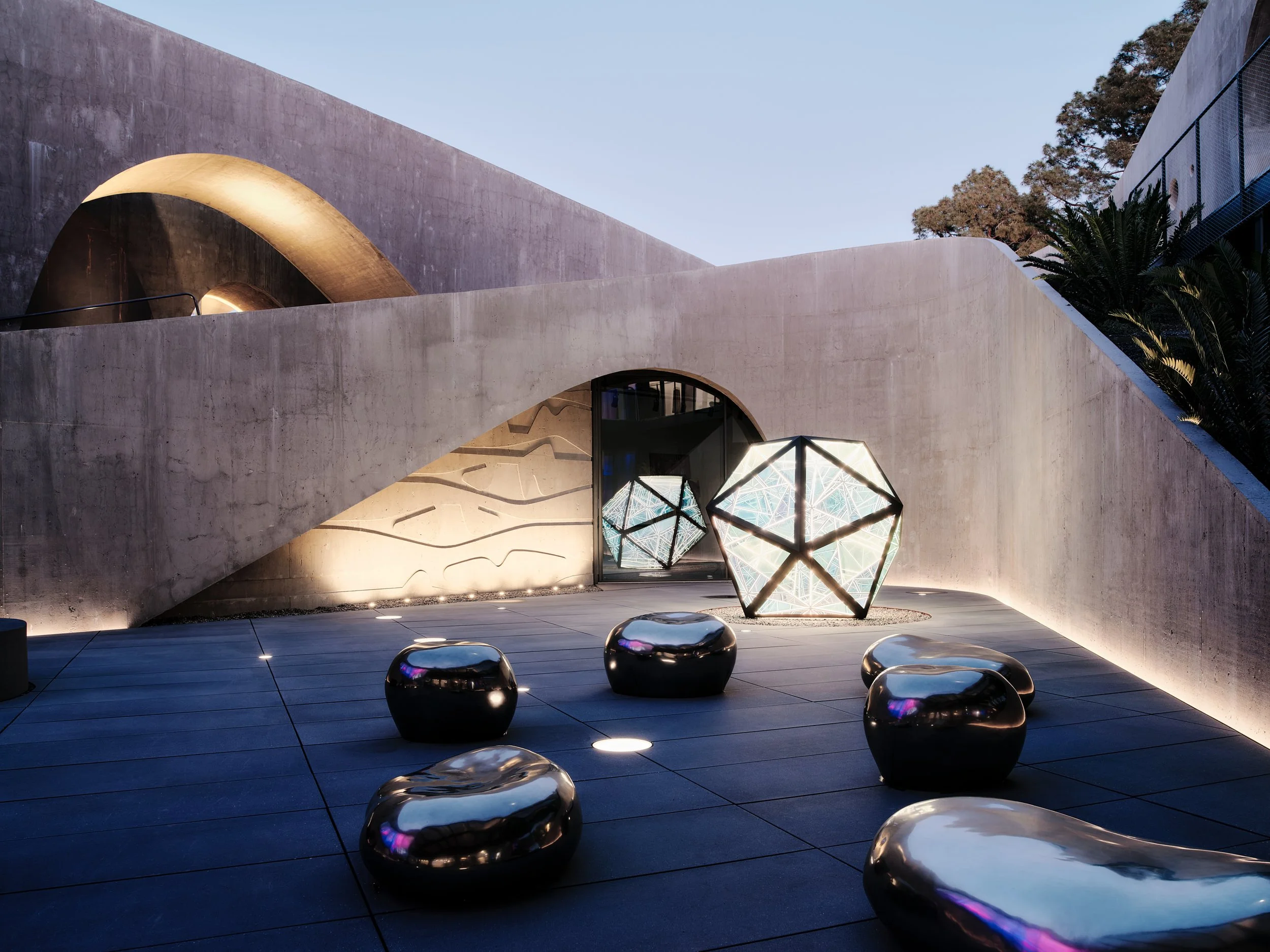 Modern architectural structure with curved concrete walls, illuminated geometric sculpture, and reflective metal seating on a tiled outdoor area during dusk.
