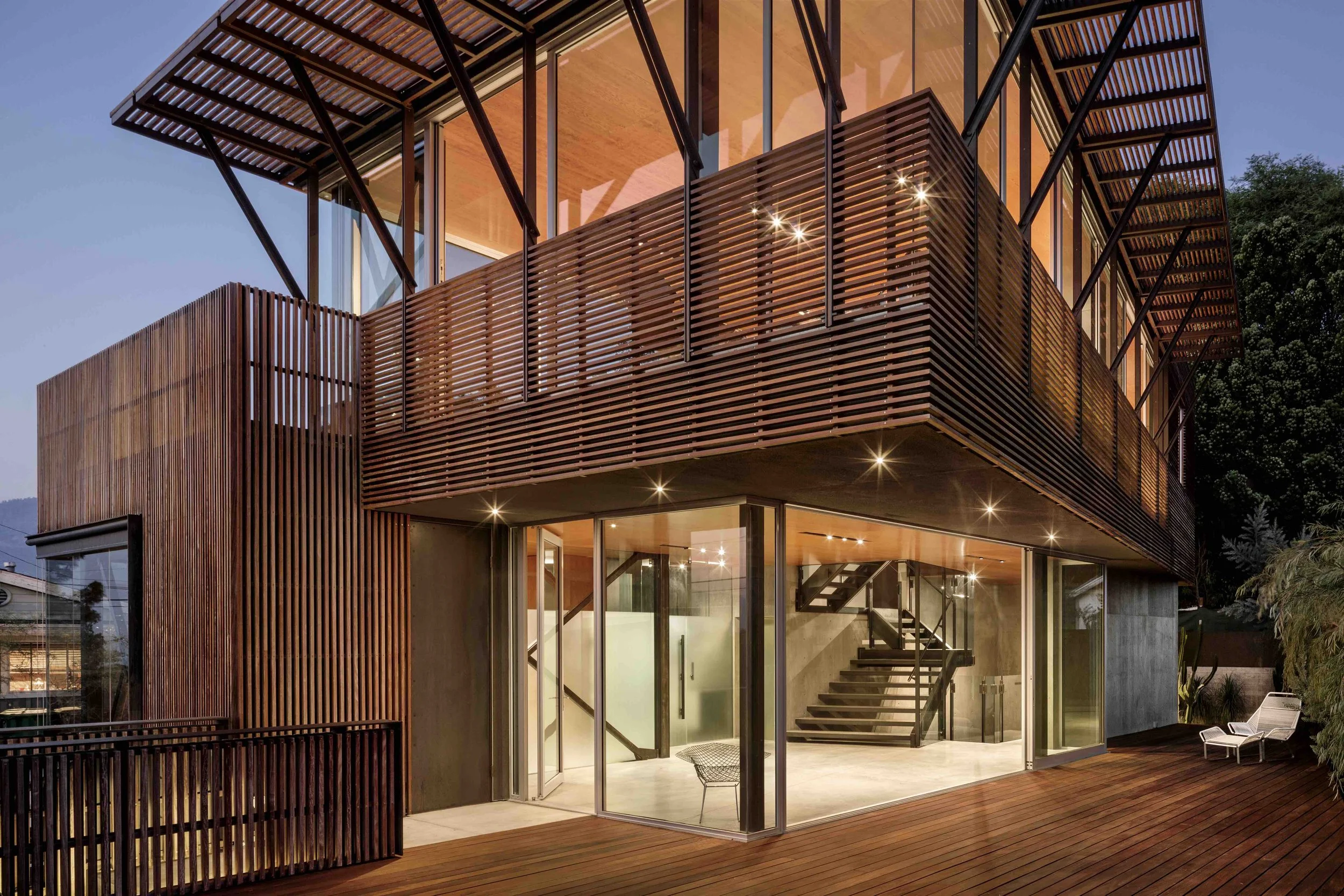 Modern multi-story house with a wooden exterior and large glass windows, illuminated from inside during the evening, surrounded by a wooden deck and greenery.