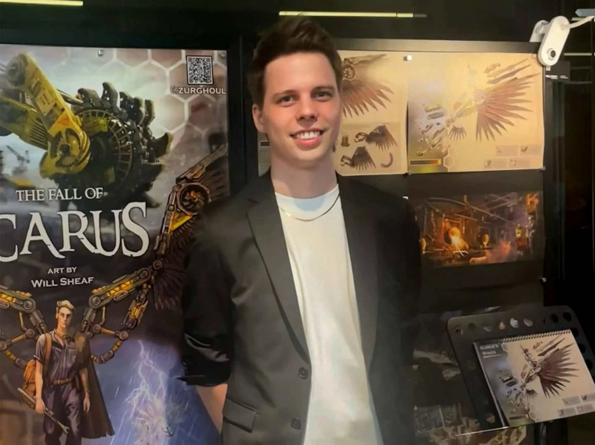 Young man smiling, standing in front of art posters and displays, including artwork titled 'The Fall of Icarus' by Will Sheaf.