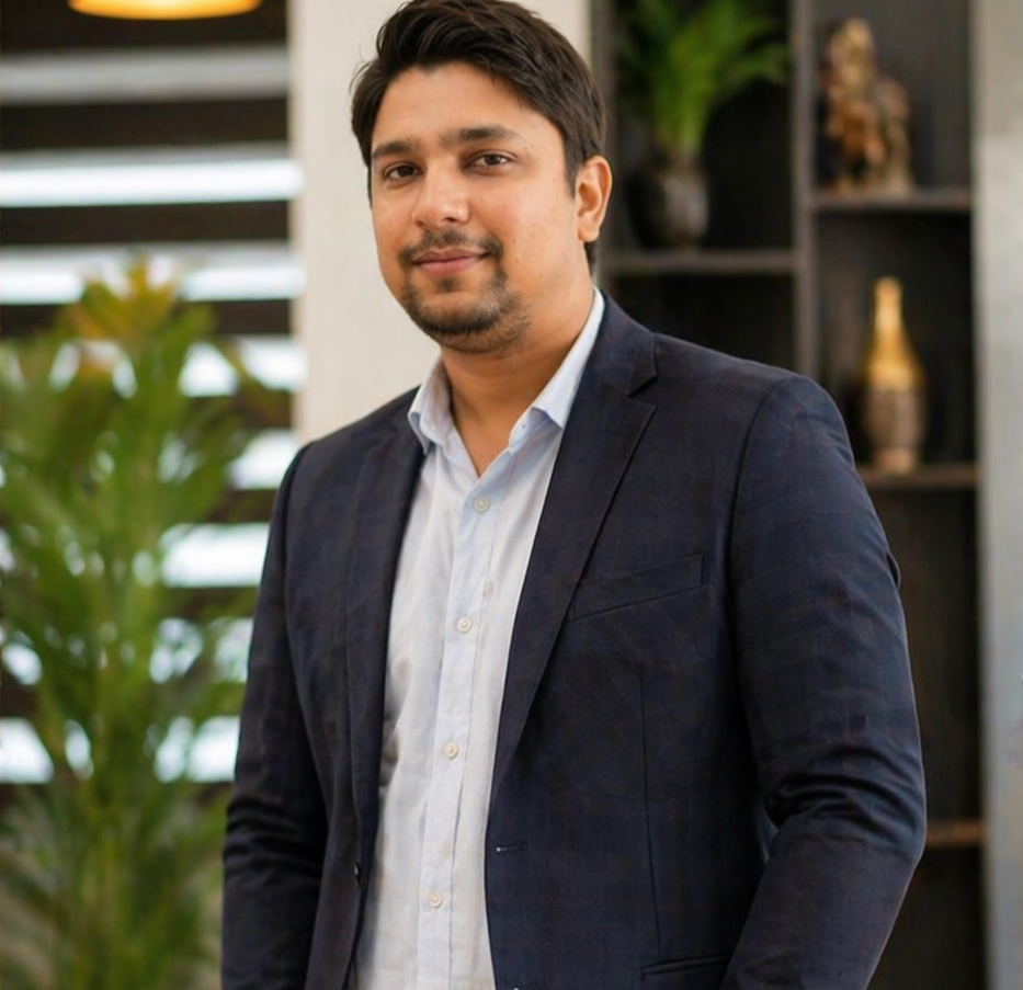 A man in a navy blazer and white shirt standing indoors with bookshelves and a plant in the background.