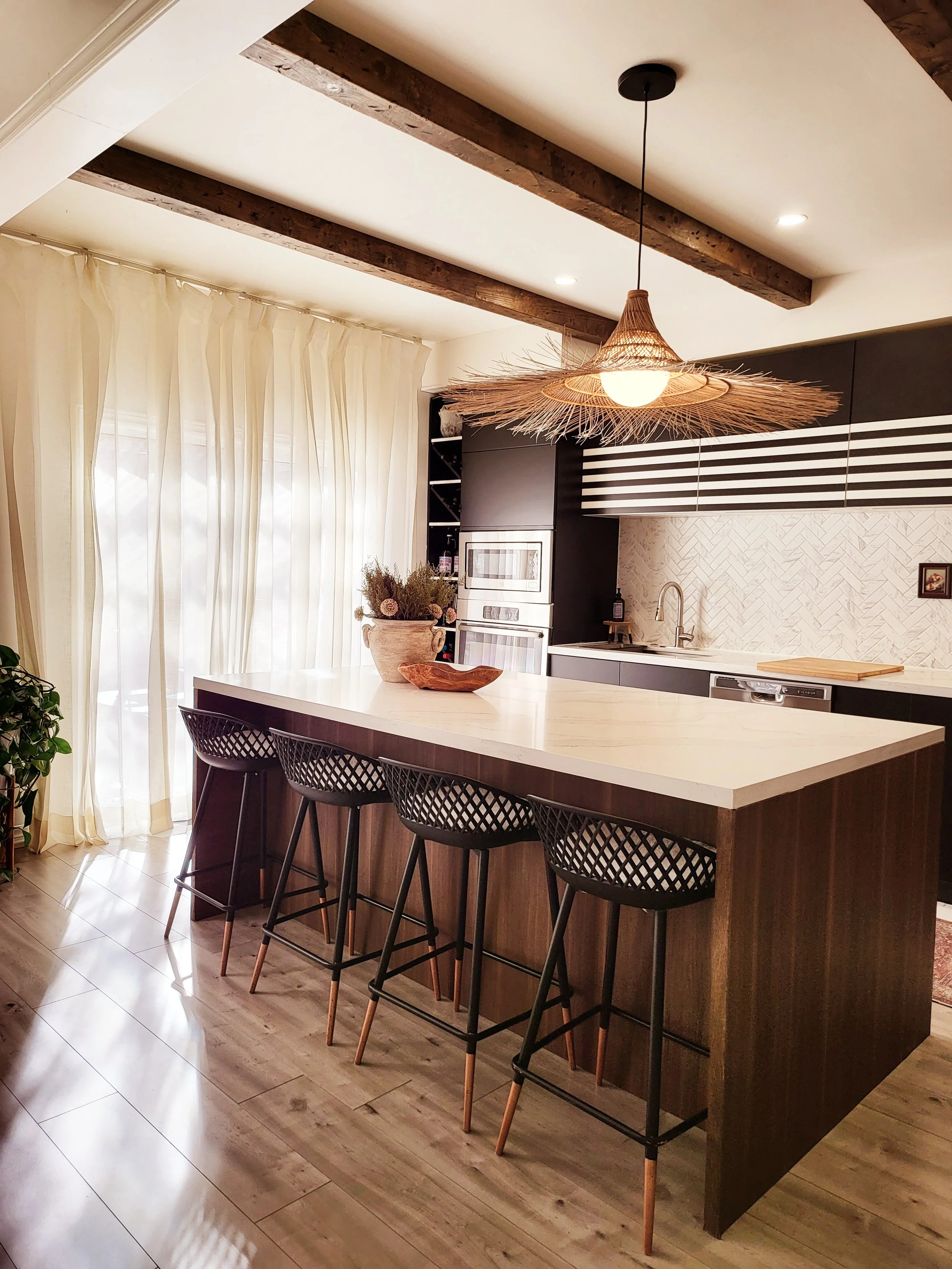 A modern kitchen with a large white island counter and four black bar stools with wooden legs. The kitchen has black cabinetry, a white countertop, and a textured white backsplash. A stylish hanging light fixture made of woven material hangs above the island. To the left, there are large cream-colored curtains covering windows, and a potted plant and decorative bowl are on the island.