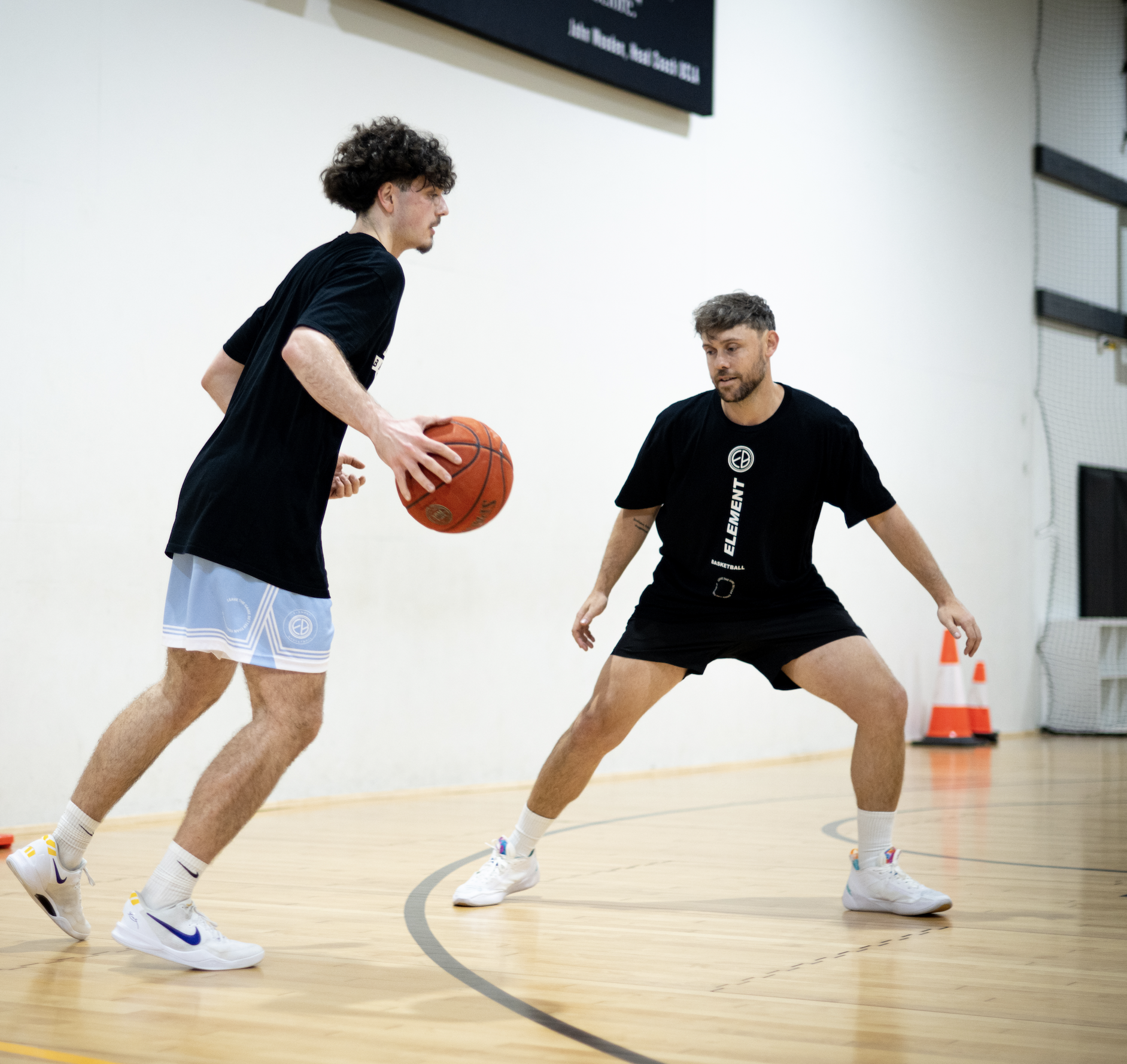 Two men playing basketball indoors, one is holding a basketball while the other is defending him, both wearing sports attire.