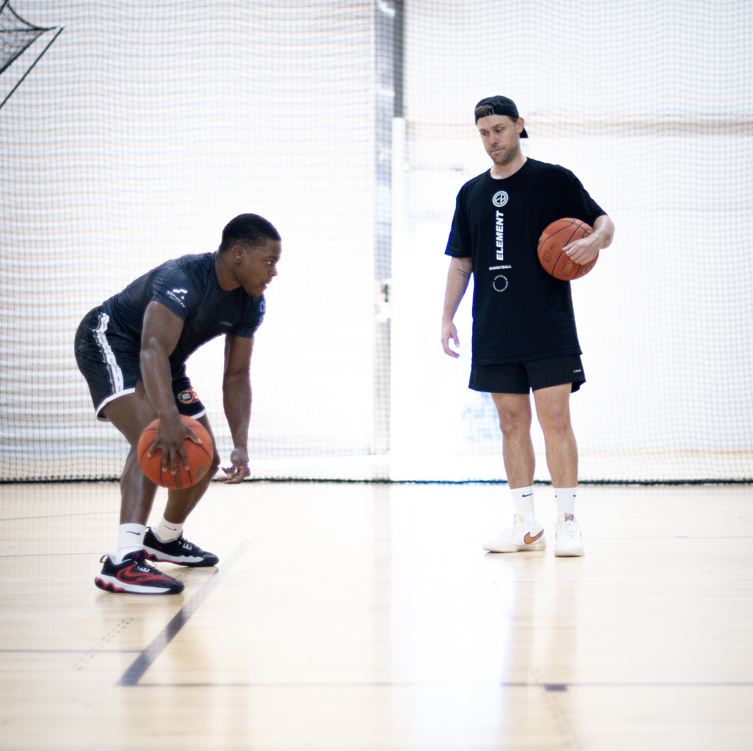 Two men playing basketball in an indoor court, one is dribbling the ball while the other observes.