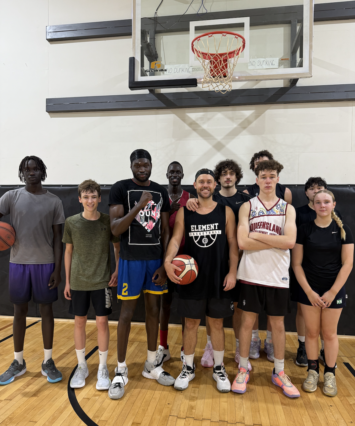 A group of young people and a coach standing on a basketball court, posing for a photo, with a basketball hoop overhead.