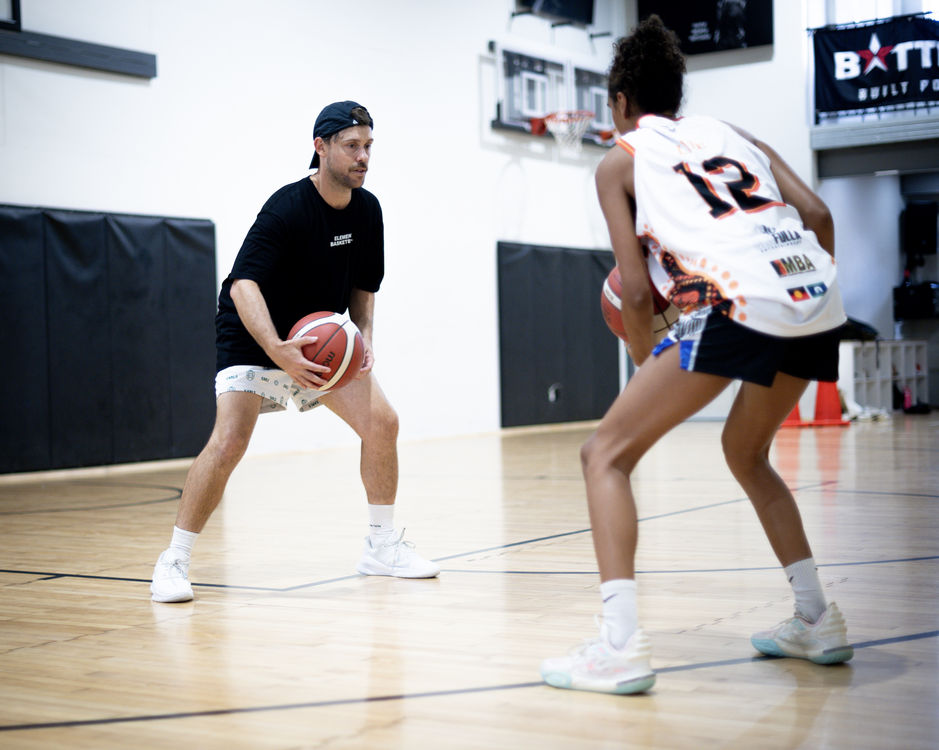 A man and a woman practicing basketball in an indoor gym. The man is holding a basketball and appears to be instructing the woman, who is in a defensive stance. Both are dressed in athletic clothing.
