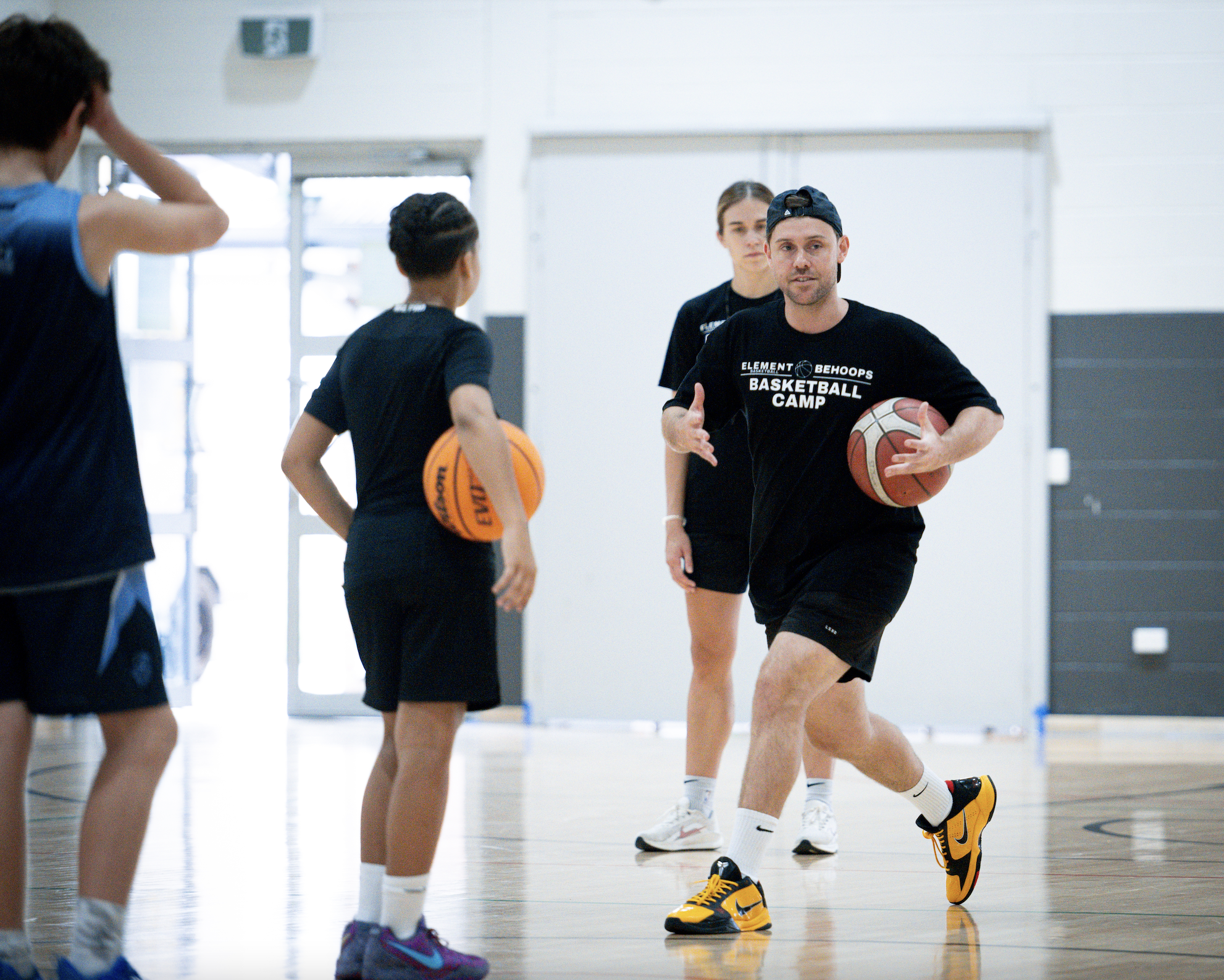 Boys and girls playing basketball in an indoor gym, with a coach instructing them.