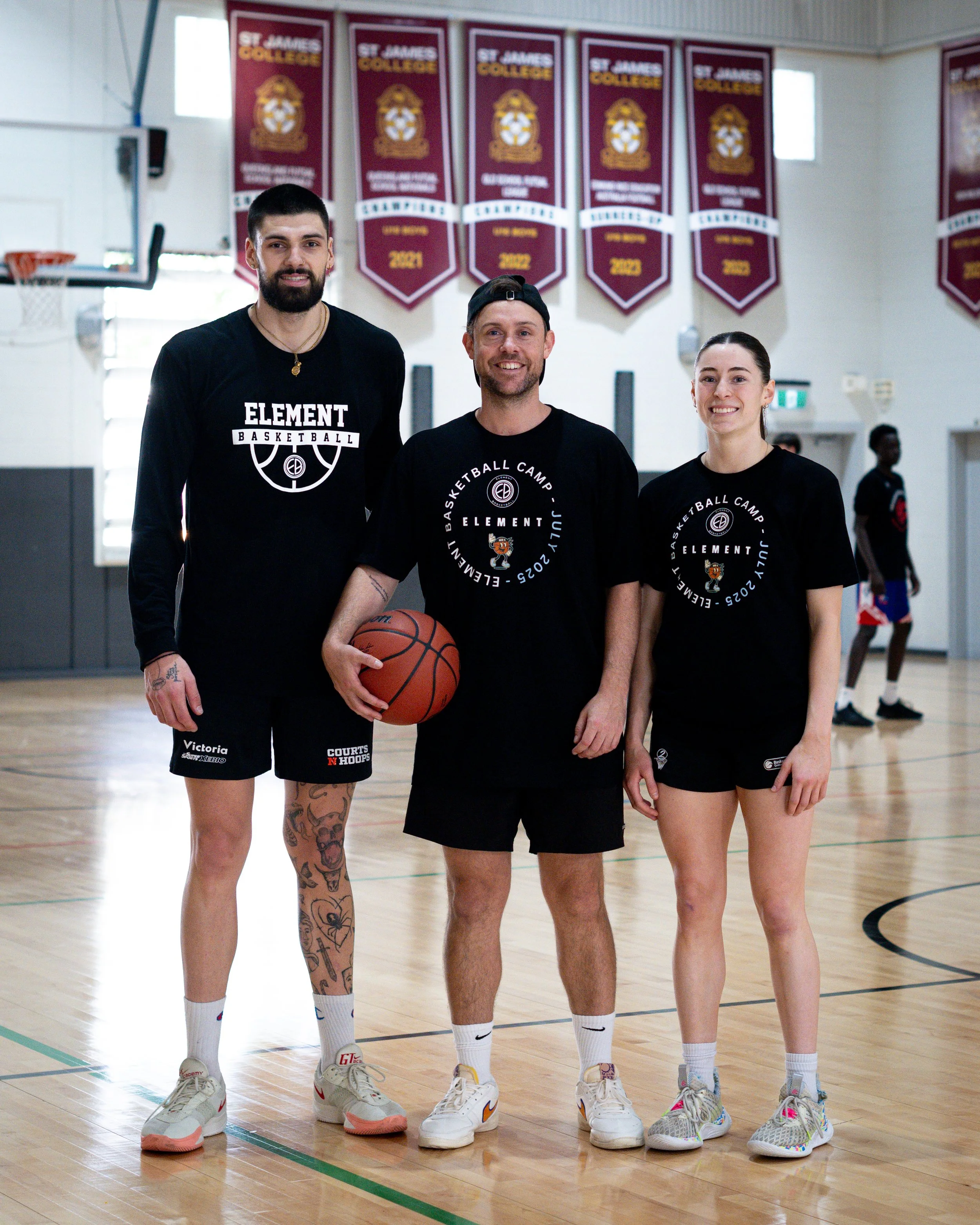 Three basketball players and a coach stand together on an indoor basketball court, smiling and posing for a photo, with banners hanging on the wall behind them.
