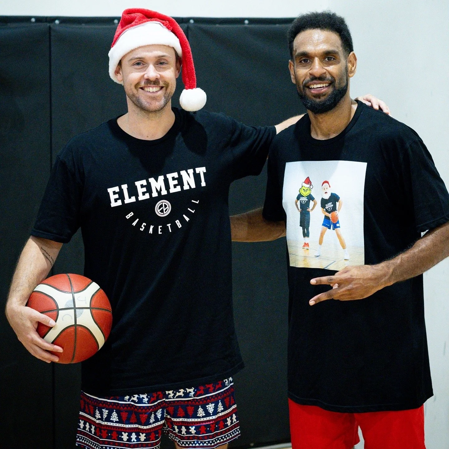 Two men standing together in a gym. The man on the left is wearing a Santa hat, a black T-shirt with "ELEMENT BASKETBALL" written on it, and patterned shorts. He is holding a basketball. The man on the right is wearing a black T-shirt with a graphic of two basketball players in Halloween costumes and red shorts. Both men are smiling and have their arms around each other.