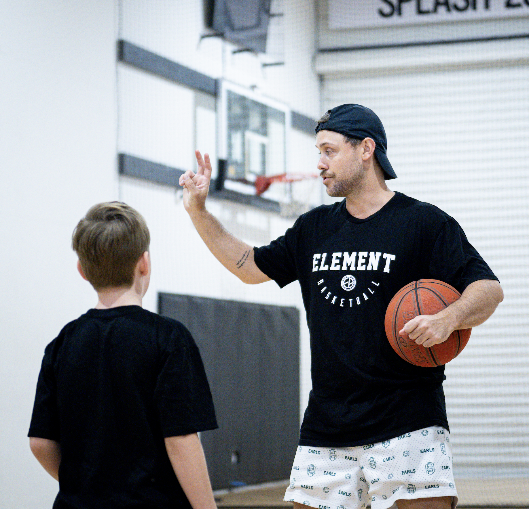 A man holding a basketball and making a gesture with his hand, talking to a young boy in a gymnasium with a basketball hoop in the background.