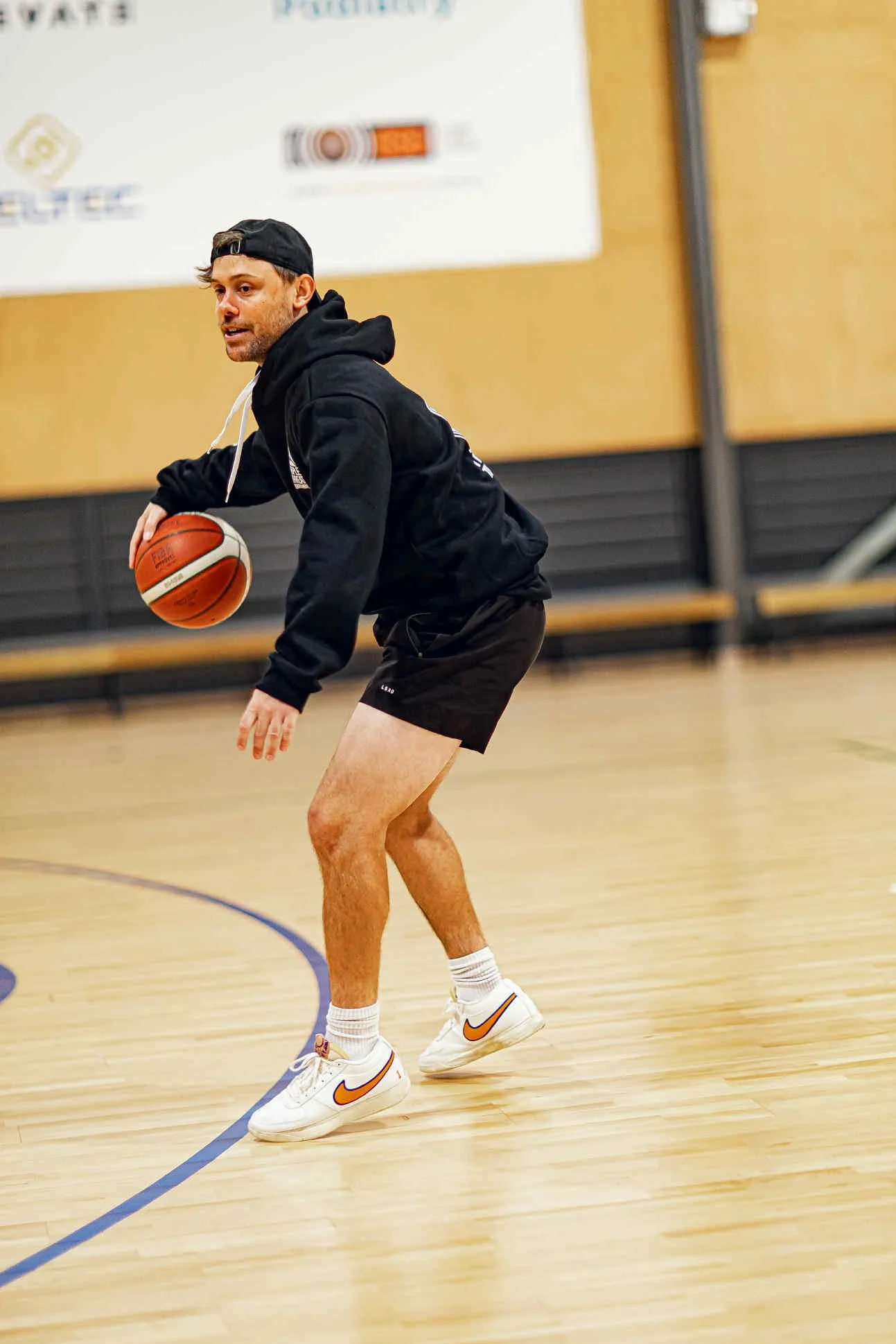 A man in a black hoodie, black shorts, and white Nike sneakers with orange swoosh, playing basketball on an indoor court.