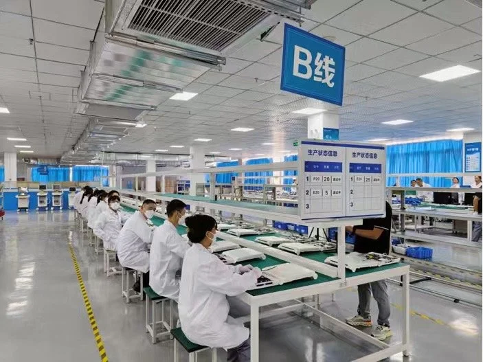 Workers wearing white lab coats and face masks sit at workstations in a large, clean manufacturing or assembly facility, with blue signs and partitions dividing sections.
