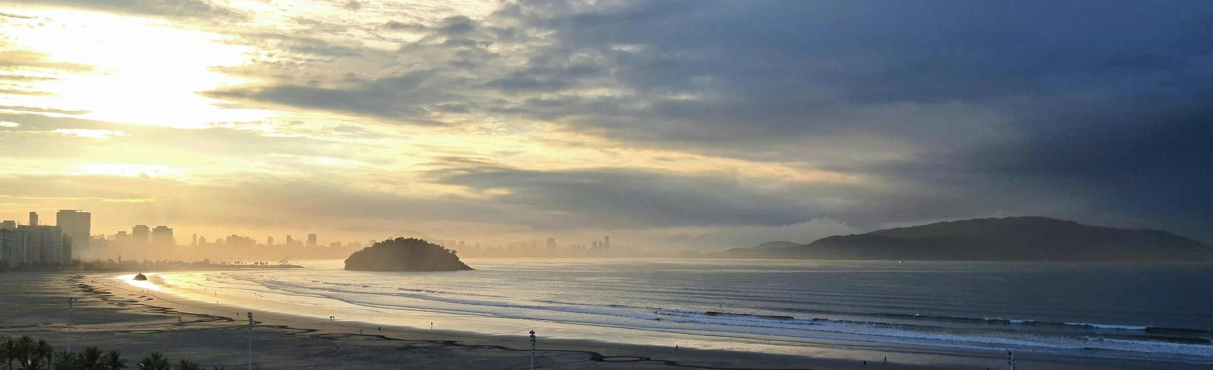Sunset over a beach with city skyline and distant hills in the background, with a partly cloudy sky.