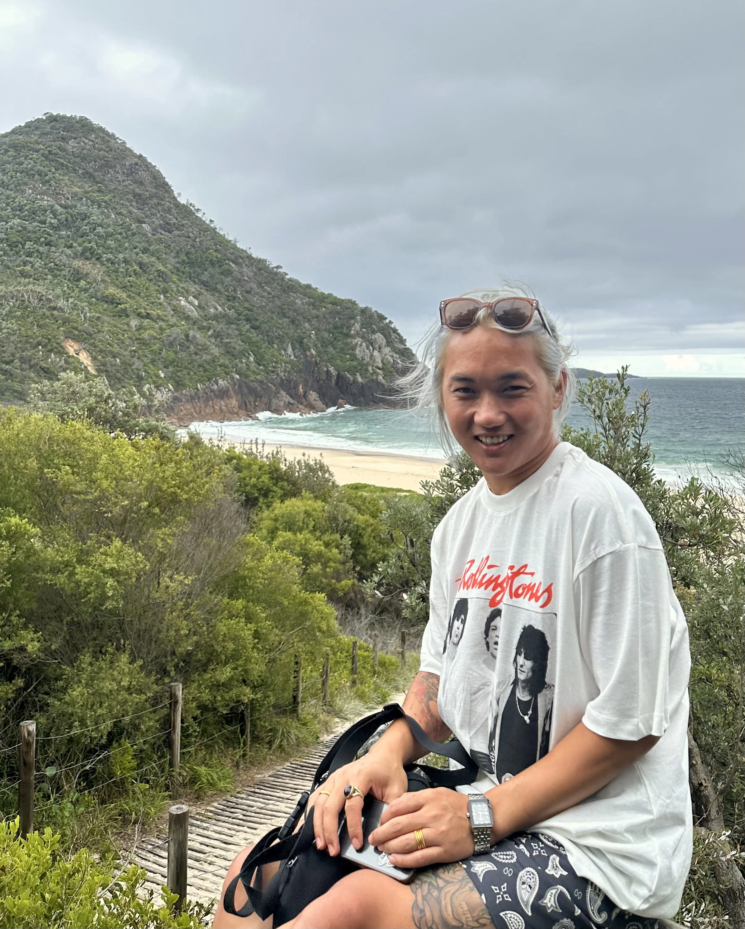 The artist Seong, wearing sunglasses on her head, a Rolling Stones t-shirt, and patterned shorts, sitting on a trail near a beach with green bushes and a mountain in the background. The ocean and cloudy sky are visible in the distance.