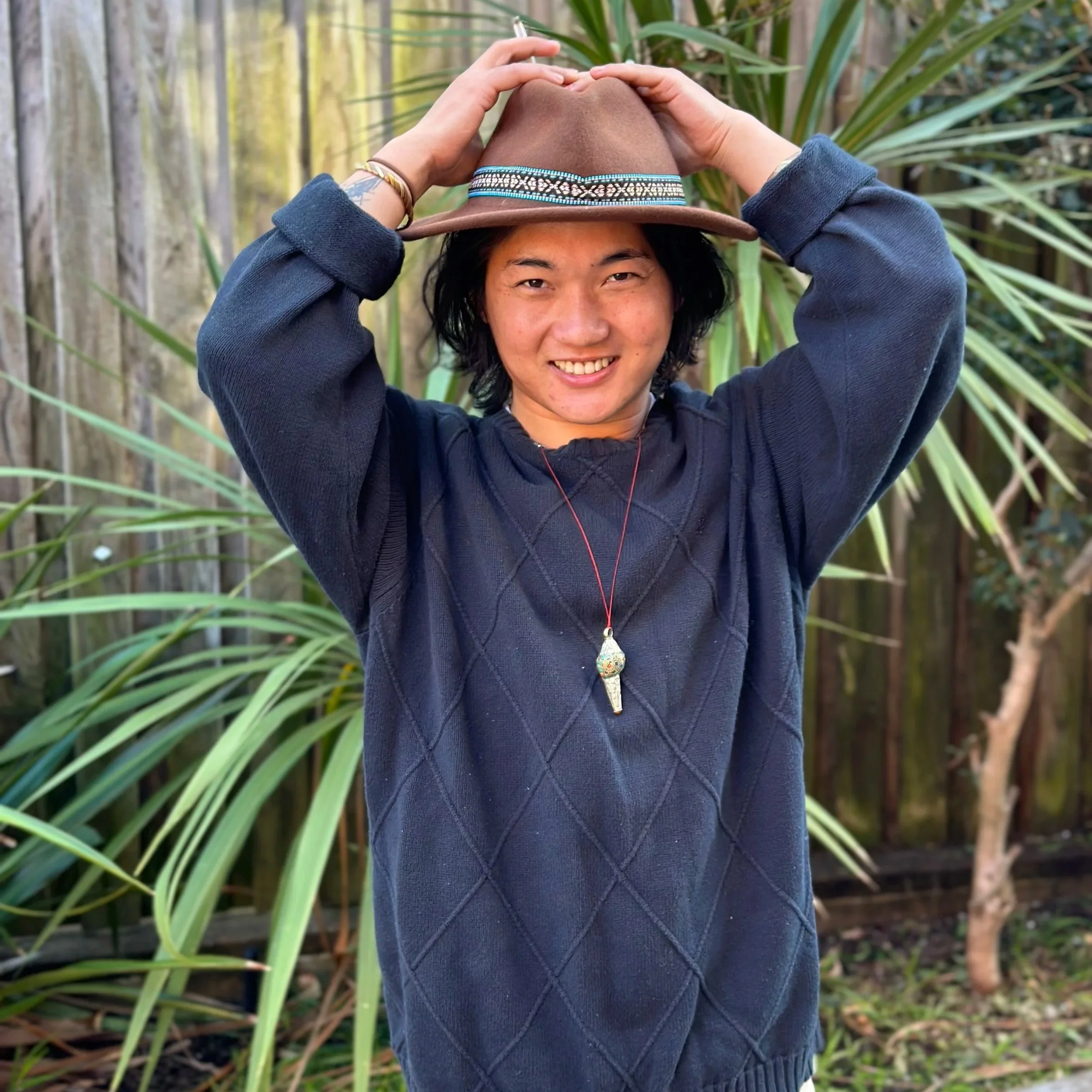 A person with shoulder-length dark hair wearing a blue sweater and a brown hat with a patterned band, smiling and adjusting their hat with both hands, standing outdoors with green plants and a wooden fence in the background.