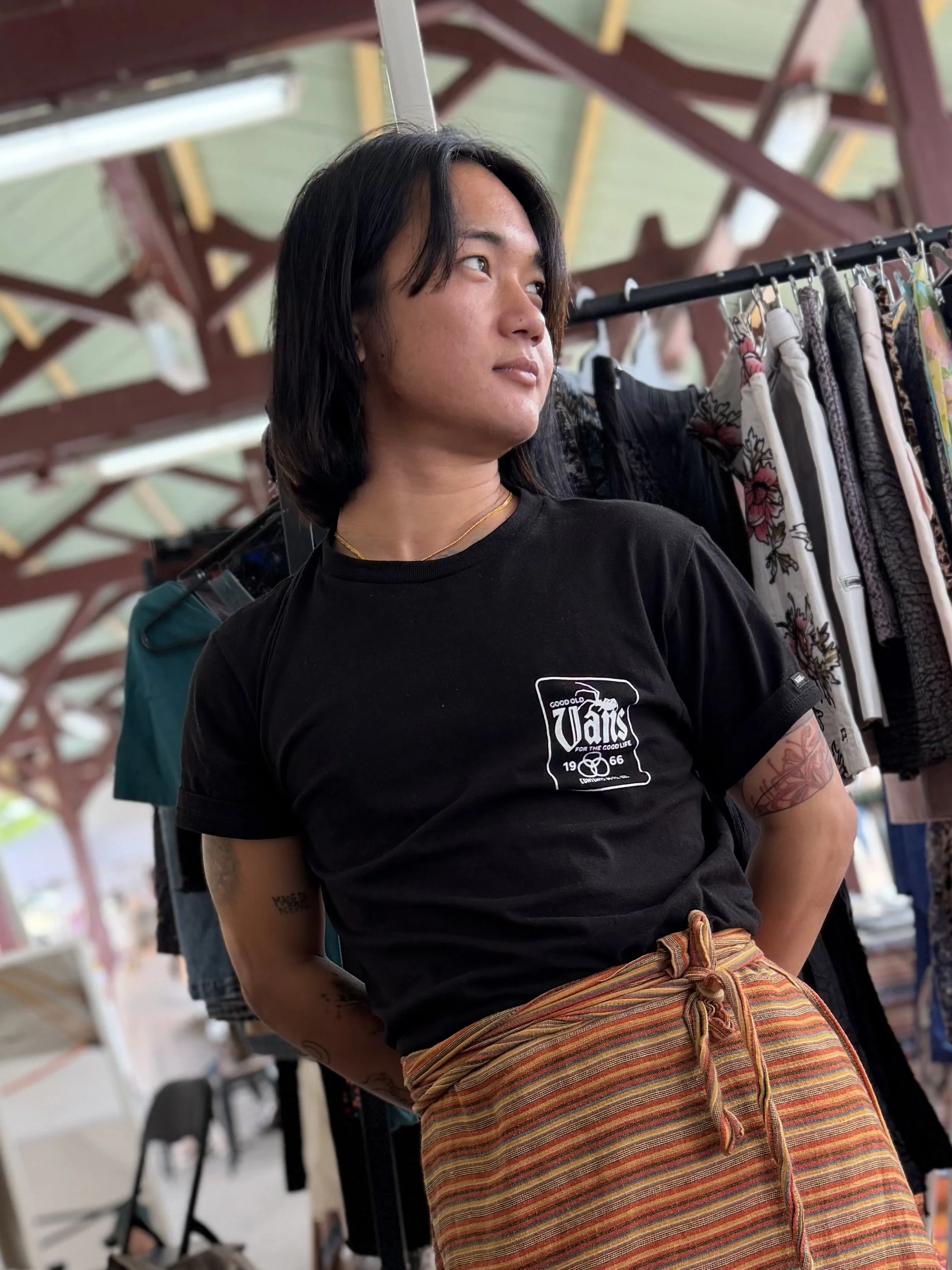 A person with medium-length dark hair wearing a black T-shirt with a white graphic design on the chest, a gold necklace, and colorful striped pants, standing near a clothing rack with various patterned garments inside a covered outdoor market or shopping area.