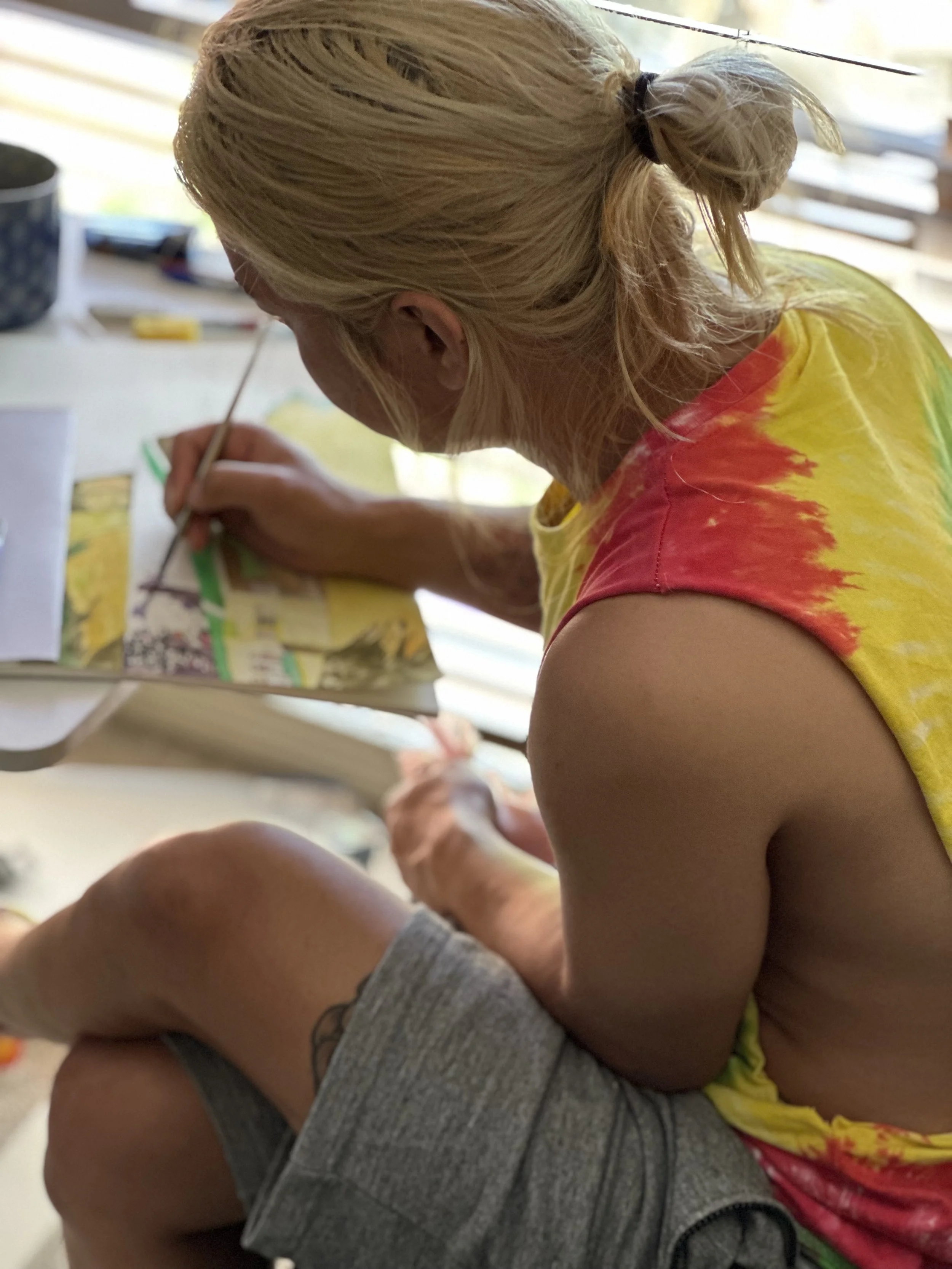 A woman with blonde hair tied back and wearing a yellow tie-dye shirt and grey shorts. She is sitting at a table, painting on a canvas with a brush, with her head bent down as she focuses on her artwork.