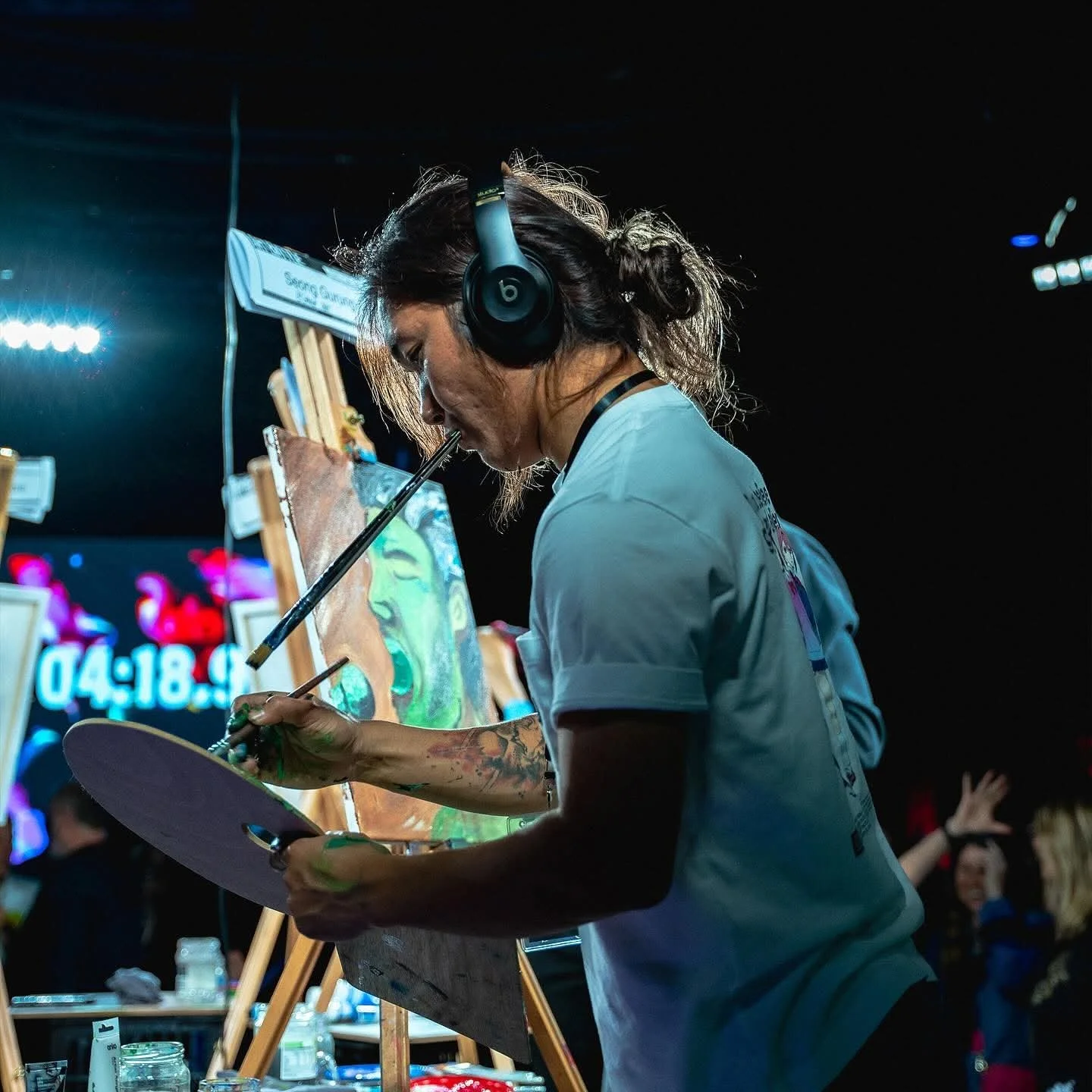 An artist painting a portrait at an event, wearing a white t-shirt and large headphones, with a timer and digital clock in the background and other people watching.