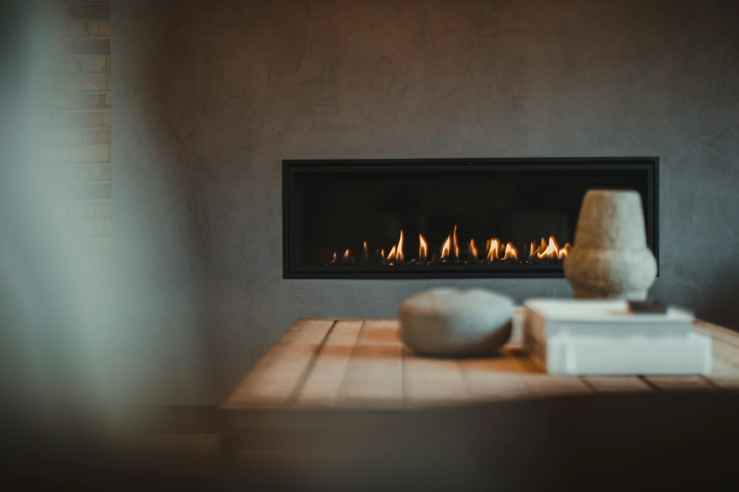 Living room with a modern fireplace with a visible flame, a wooden table with pottery and decorative objects, and a gray wall background.