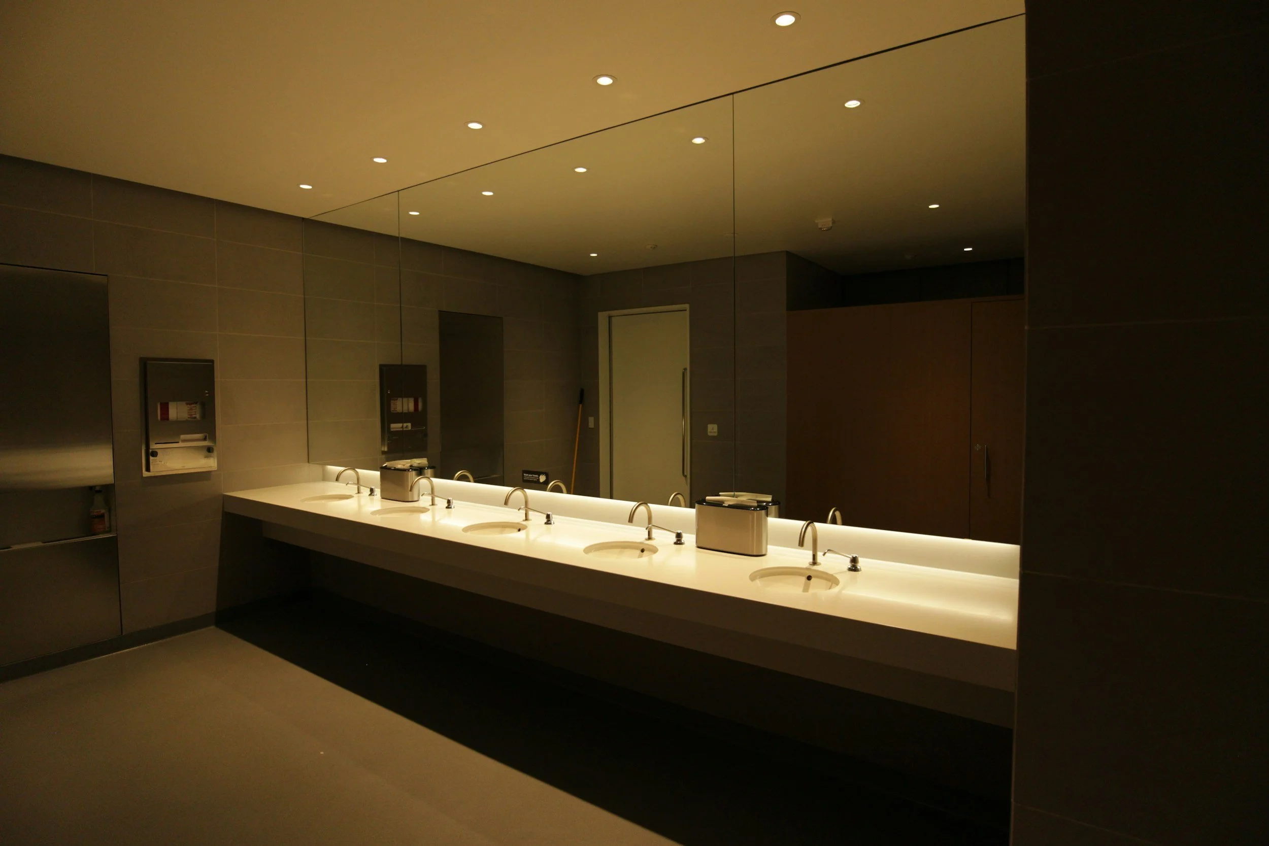 A modern restroom with a long white countertop featuring three sinks, mirrors covering the wall behind, and a ceiling with recessed lighting.