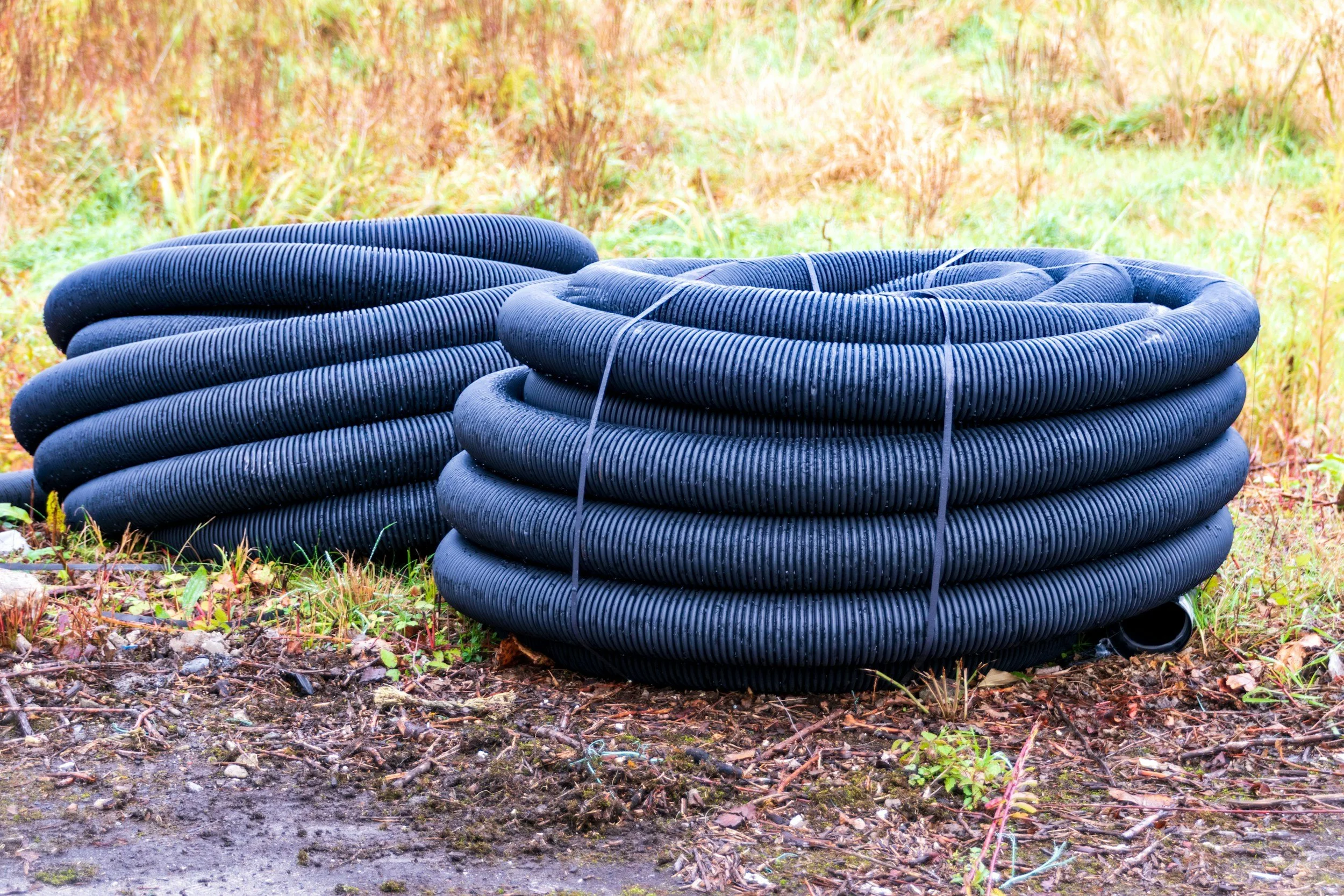 Two large coils of black corrugated pipe laid on grass and soil in an outdoor setting with green and yellow vegetation in the background.