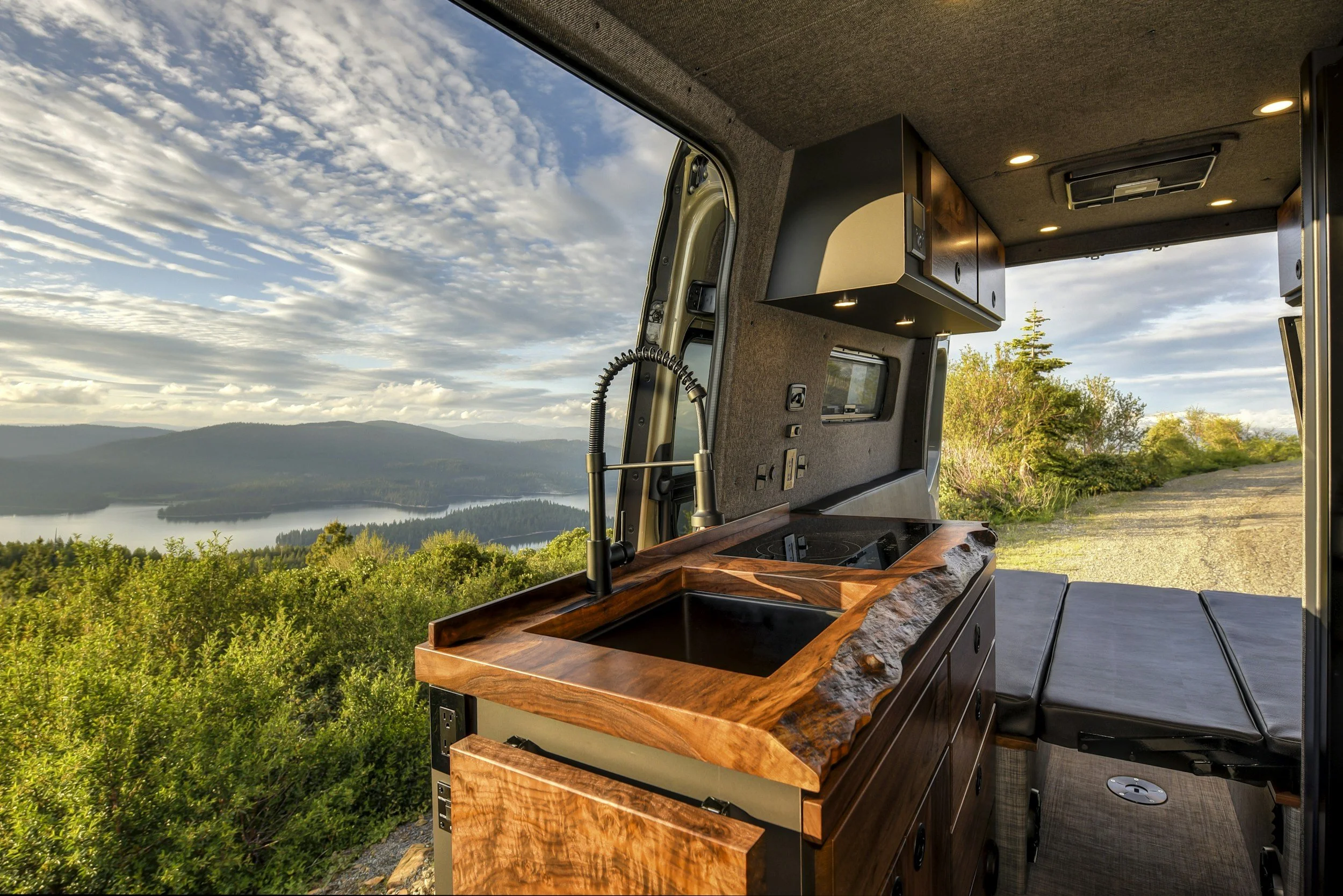 View from inside a camper van, showing a modern kitchen area with a wooden counter and sink, overlooking a scenic landscape of trees, hills, and a body of water under a partly cloudy sky.