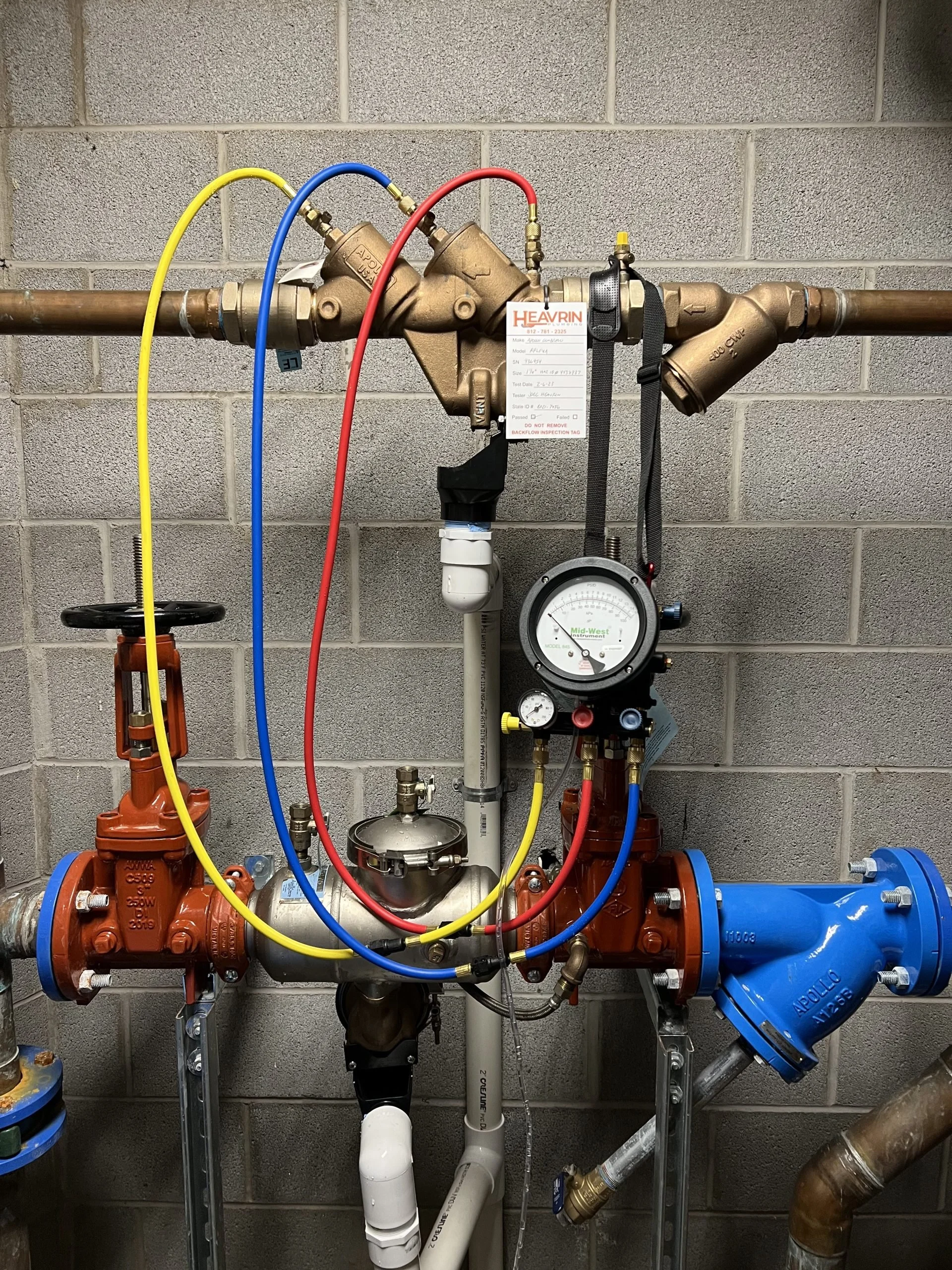 Industrial plumbing system with copper and PVC pipes, valves, gauges, and color-coded hoses mounted on a cinder block wall.