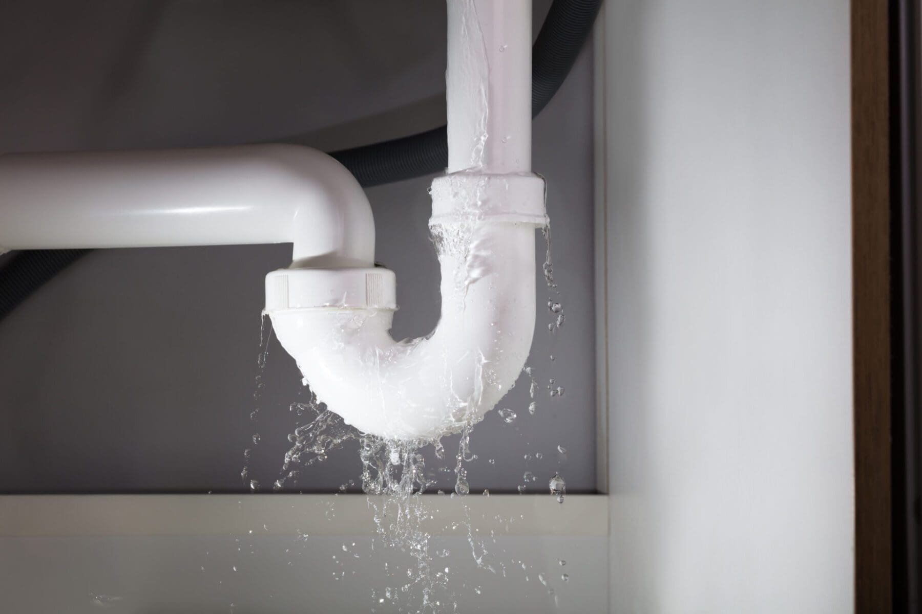 A close-up of a white PVC water pipe with water leaking and dripping from it, under a sink.
