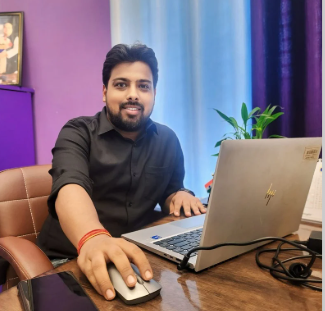 Man sitting at desk with laptop, mouse, and potted plant in a colorful room with purple and blue curtains.