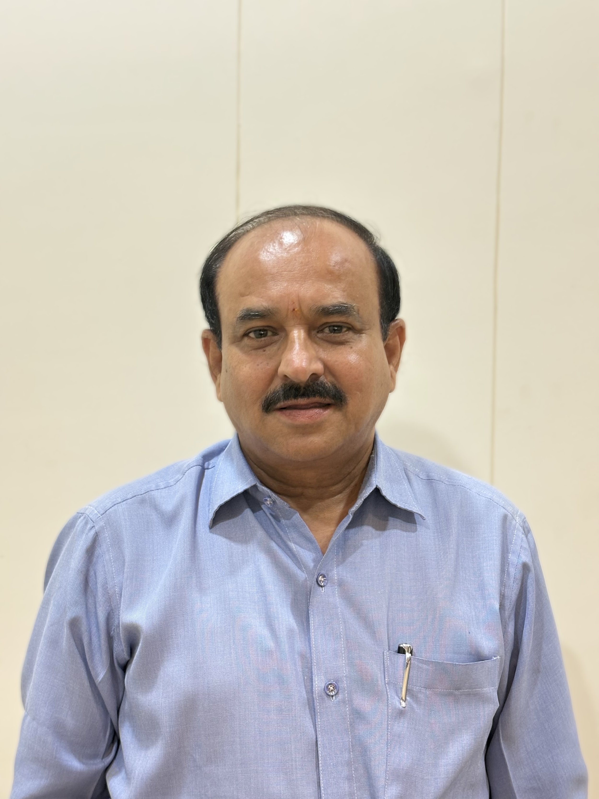 A man with a mustache wearing a light blue button-up shirt standing in front of a plain beige wall.