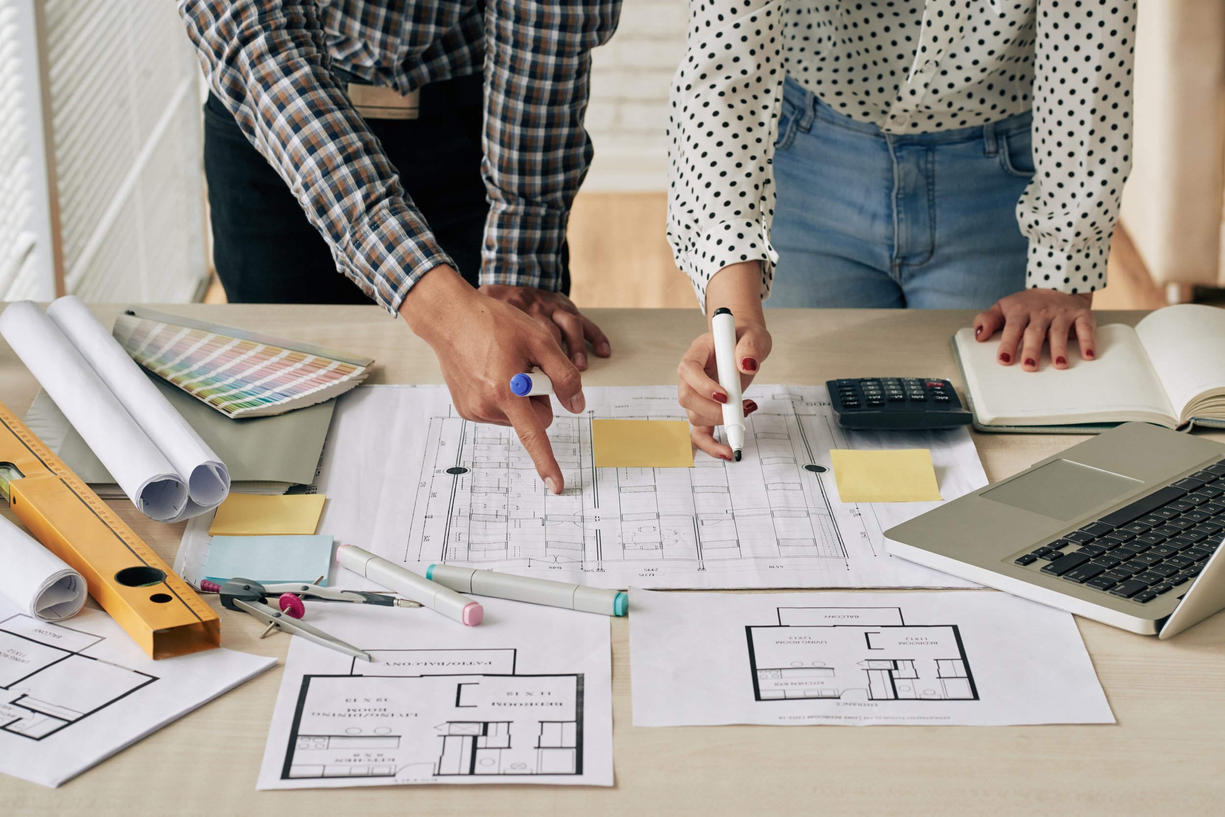 Two people working on architectural blueprints and plans, with office supplies and a laptop on the table.