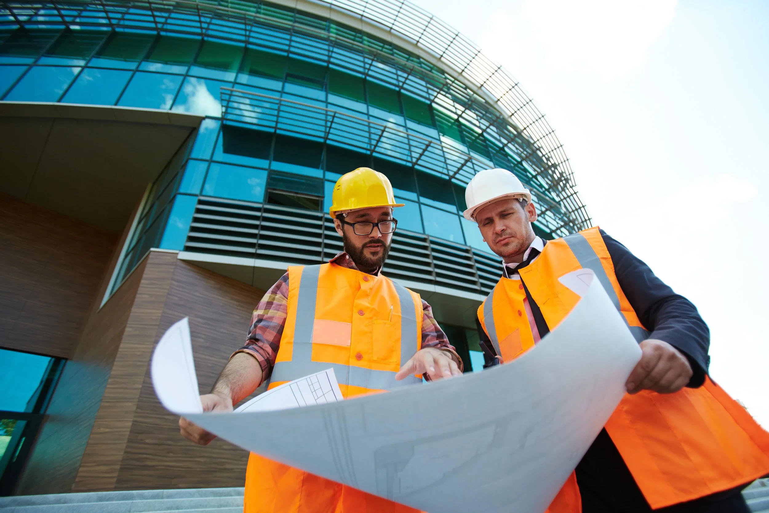Two construction workers wearing orange safety vests and hard hats reviewing blueprints outside a modern glass building.