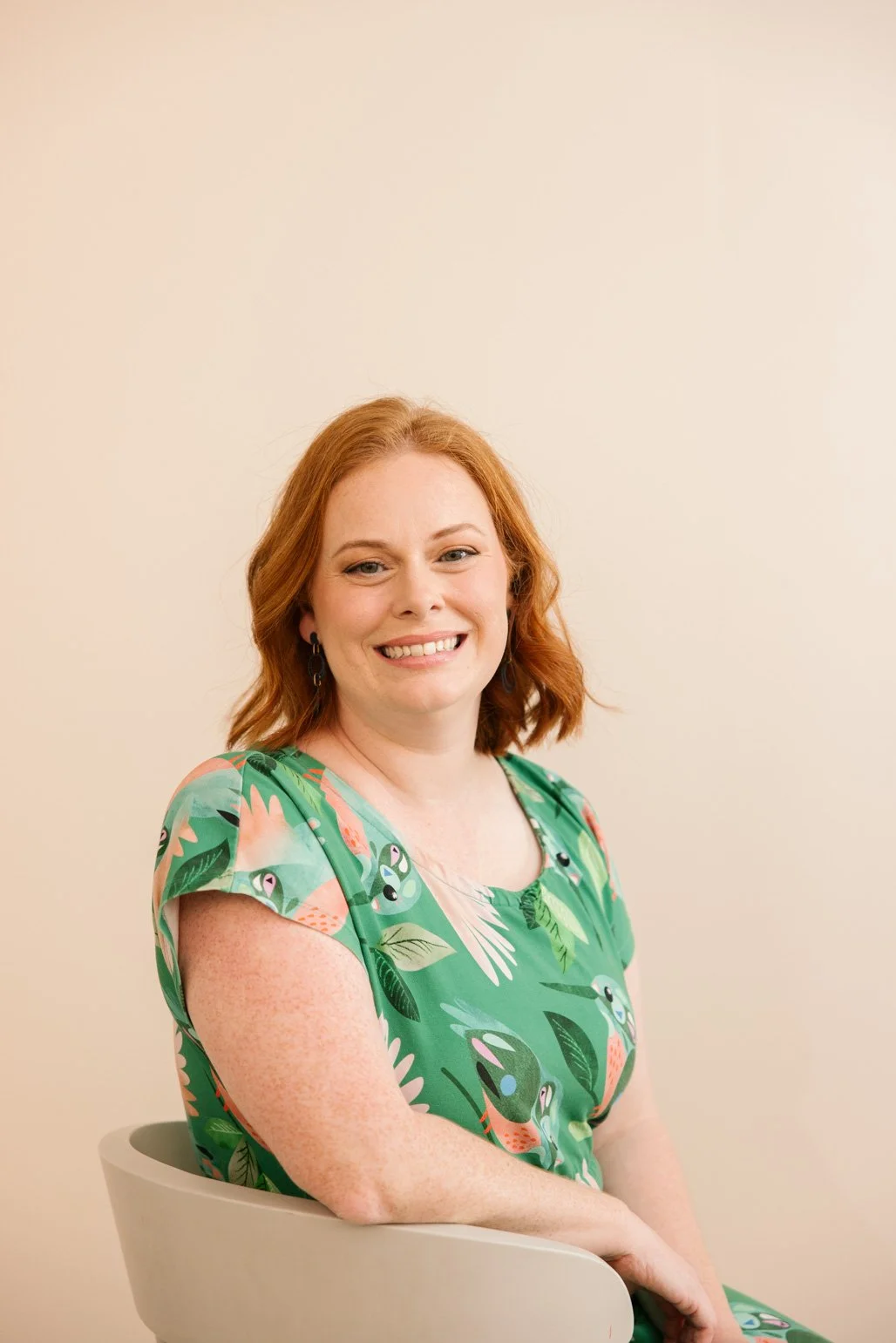 Mel Williamson, Trauma therapist,  wearing a green dress with tropical bird and leaf patterns, sitting on a light grey chair, smiling at the camera against a plain off-white wall.
