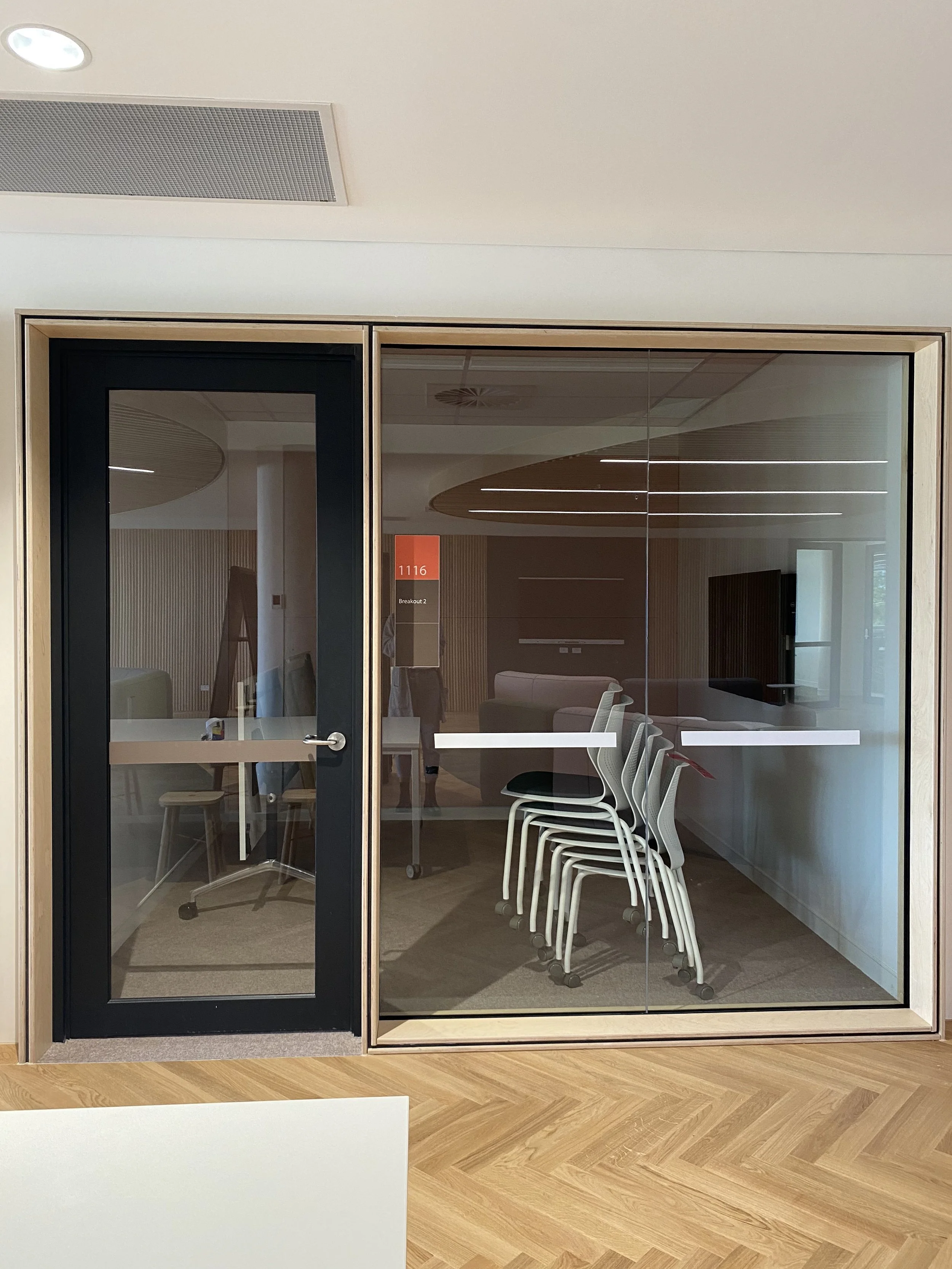 Glass meeting room with stacked chairs, modern interior design, wooden floor, and a small red sign on the door indicating room number 1116.