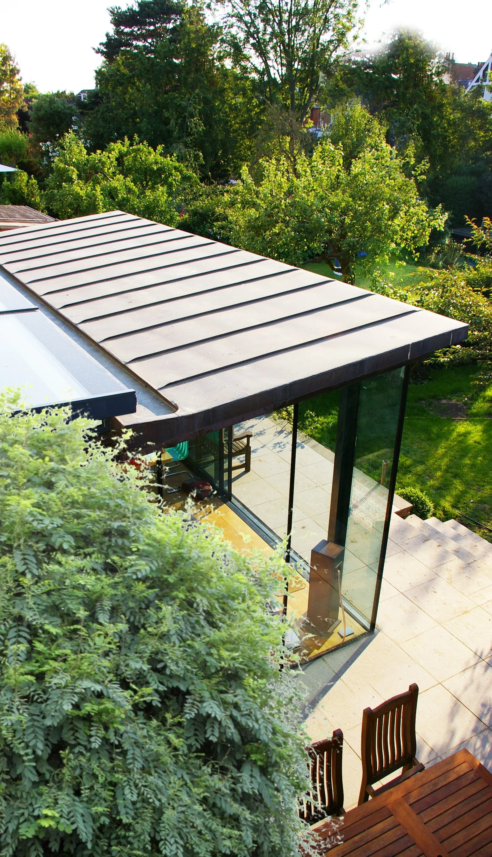 Aerial view of a modern home with a metal roof, large glass patio doors, and outdoor seating area surrounded by lush green trees and garden.
