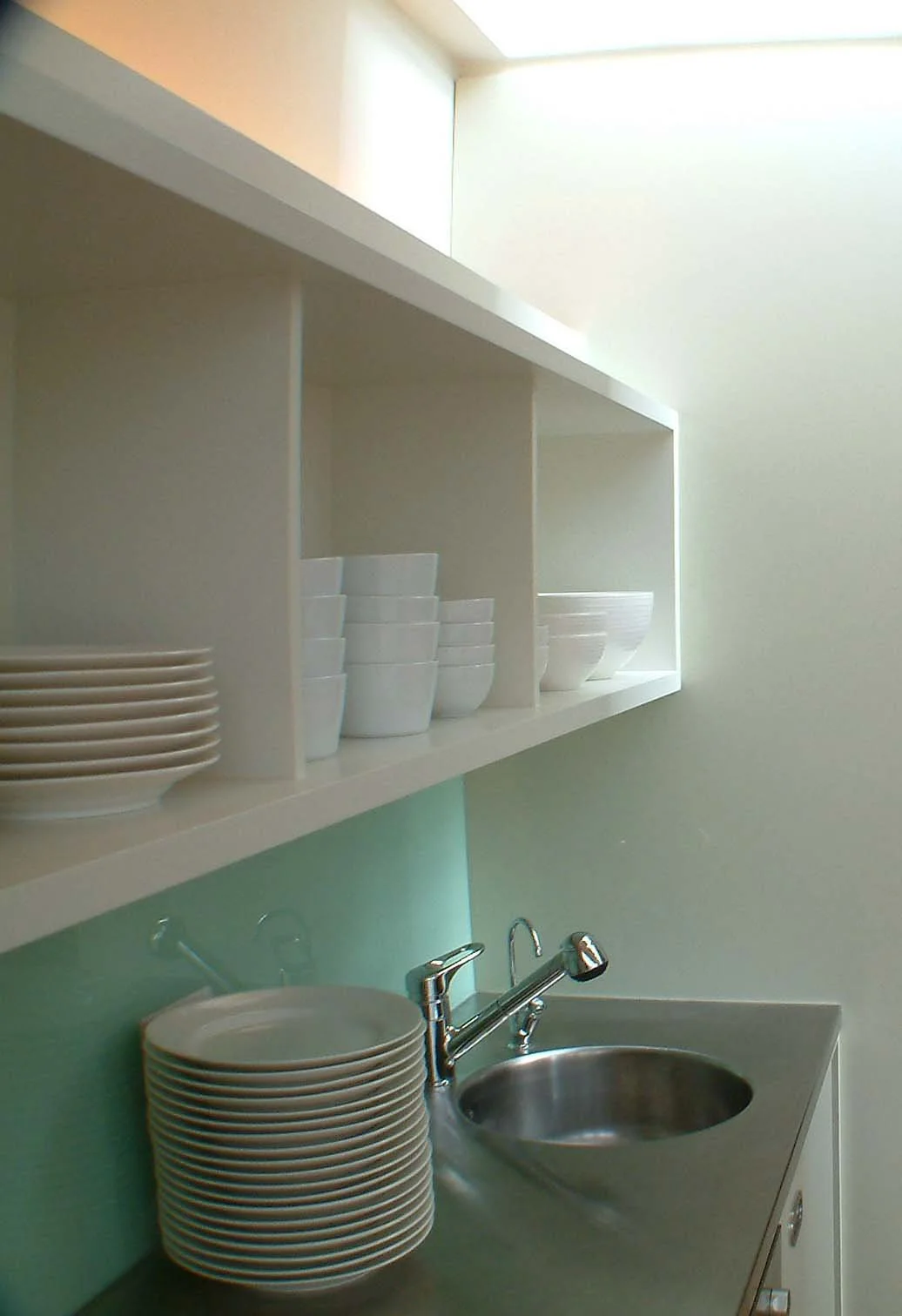 Kitchen area with open white shelves containing stacked white bowls and plates, a stainless steel sink with a chrome faucet, and a light-colored wall.