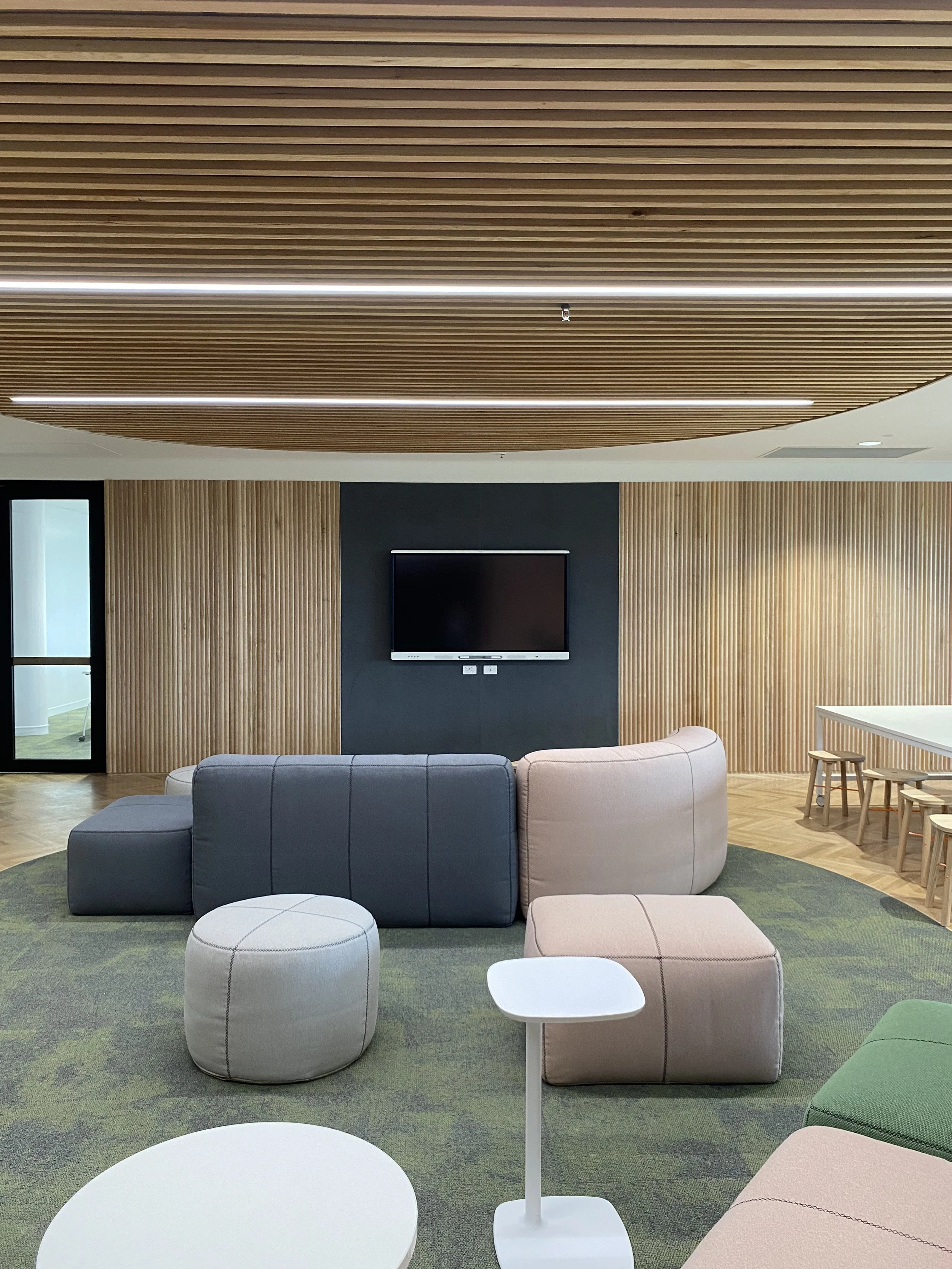 Modern lounge area with gray and pink upholstered seating, white tables, a television mounted on a dark accent wall, and wooden slat walls and ceiling panels.