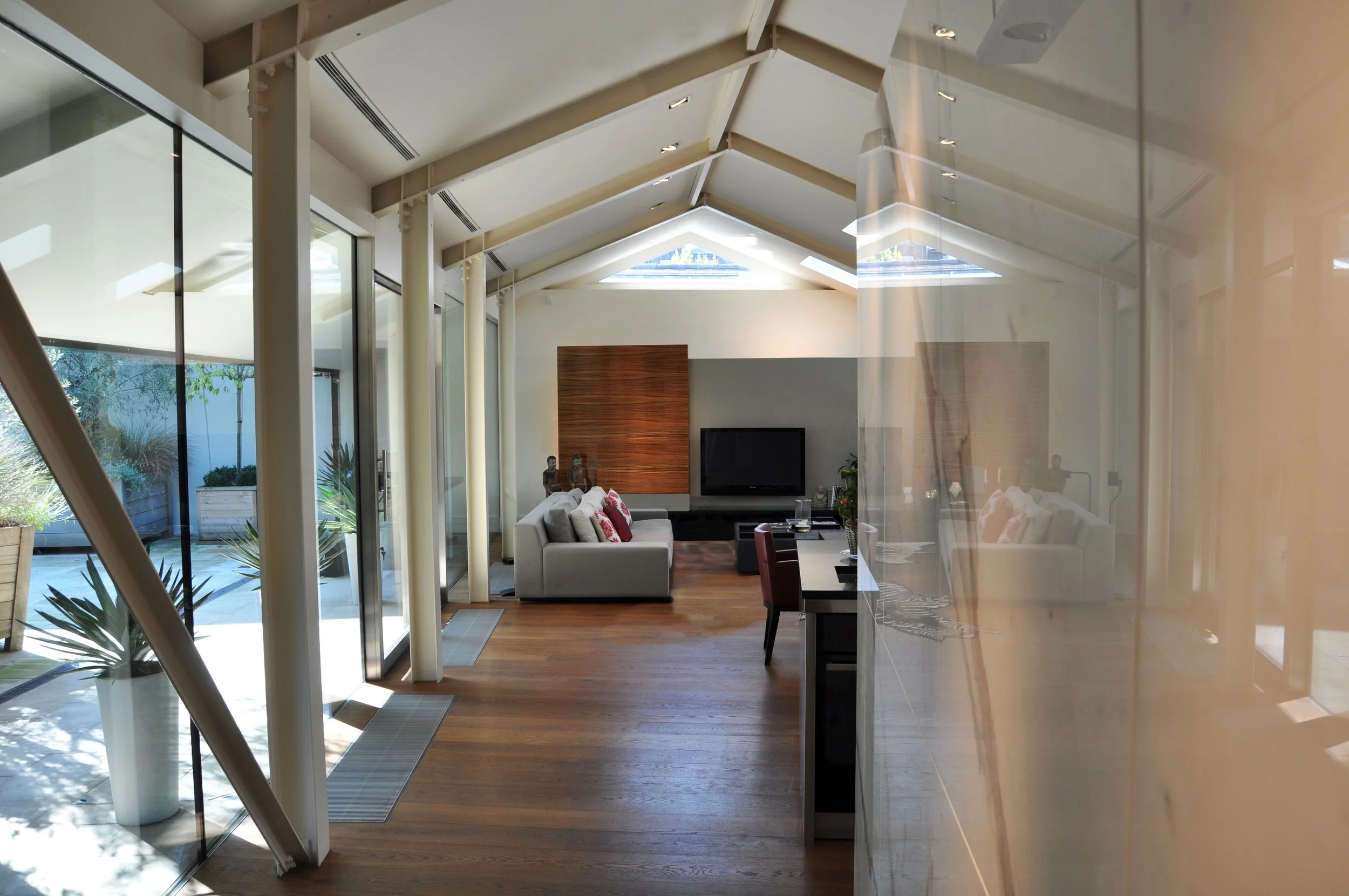 Modern open-plan living room with a sloped ceiling, large windows, and sliding glass doors leading to an outdoor patio with plants. Contains a sofa with cushions, a wooden accent wall with a flat-screen TV, and a dining table with chairs.