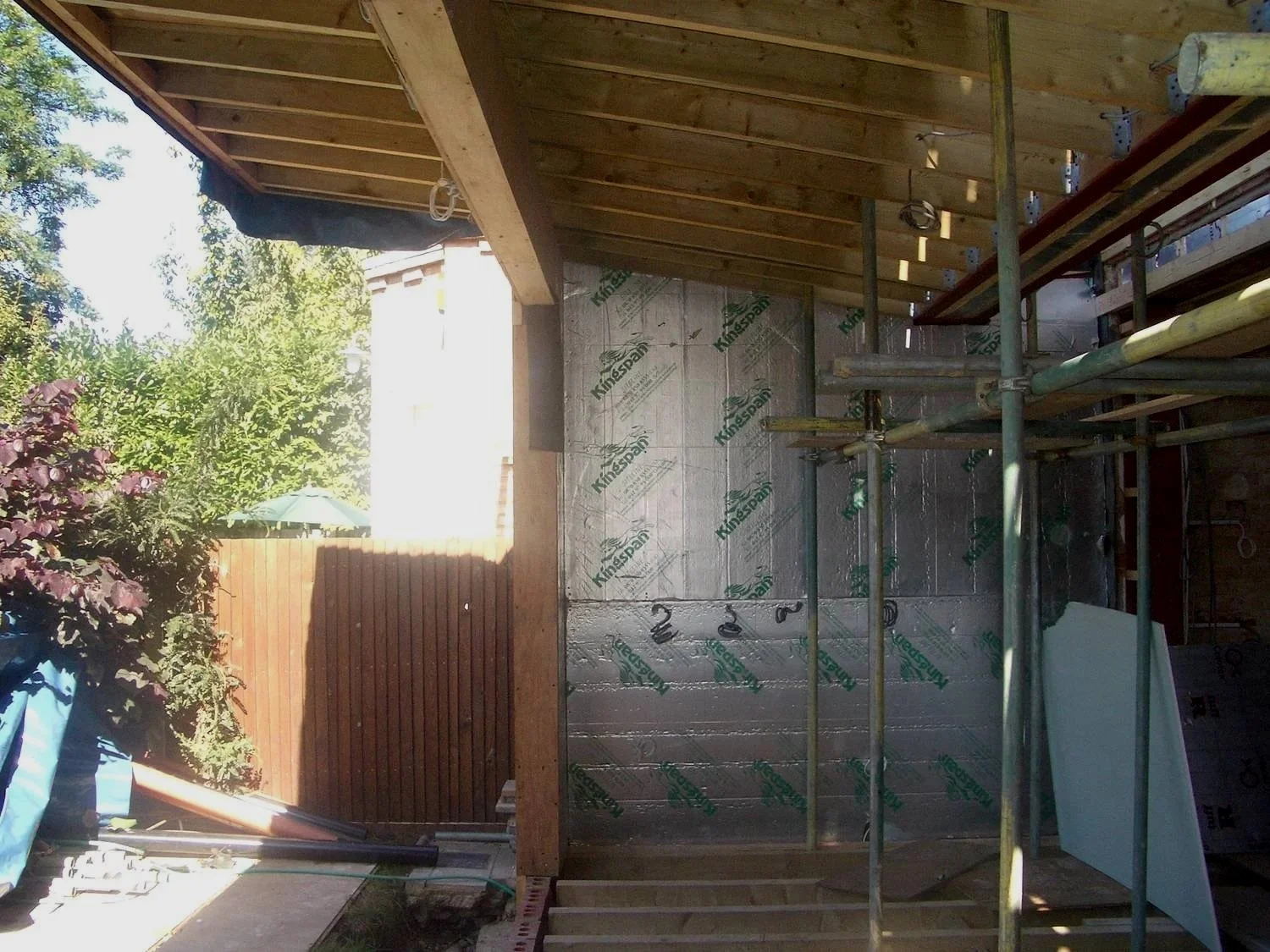 Construction of a building with wooden framing and insulation panels, scaffolding set up in the interior, and an outdoor area with a wooden fence, trees, and a patio umbrella in the background.