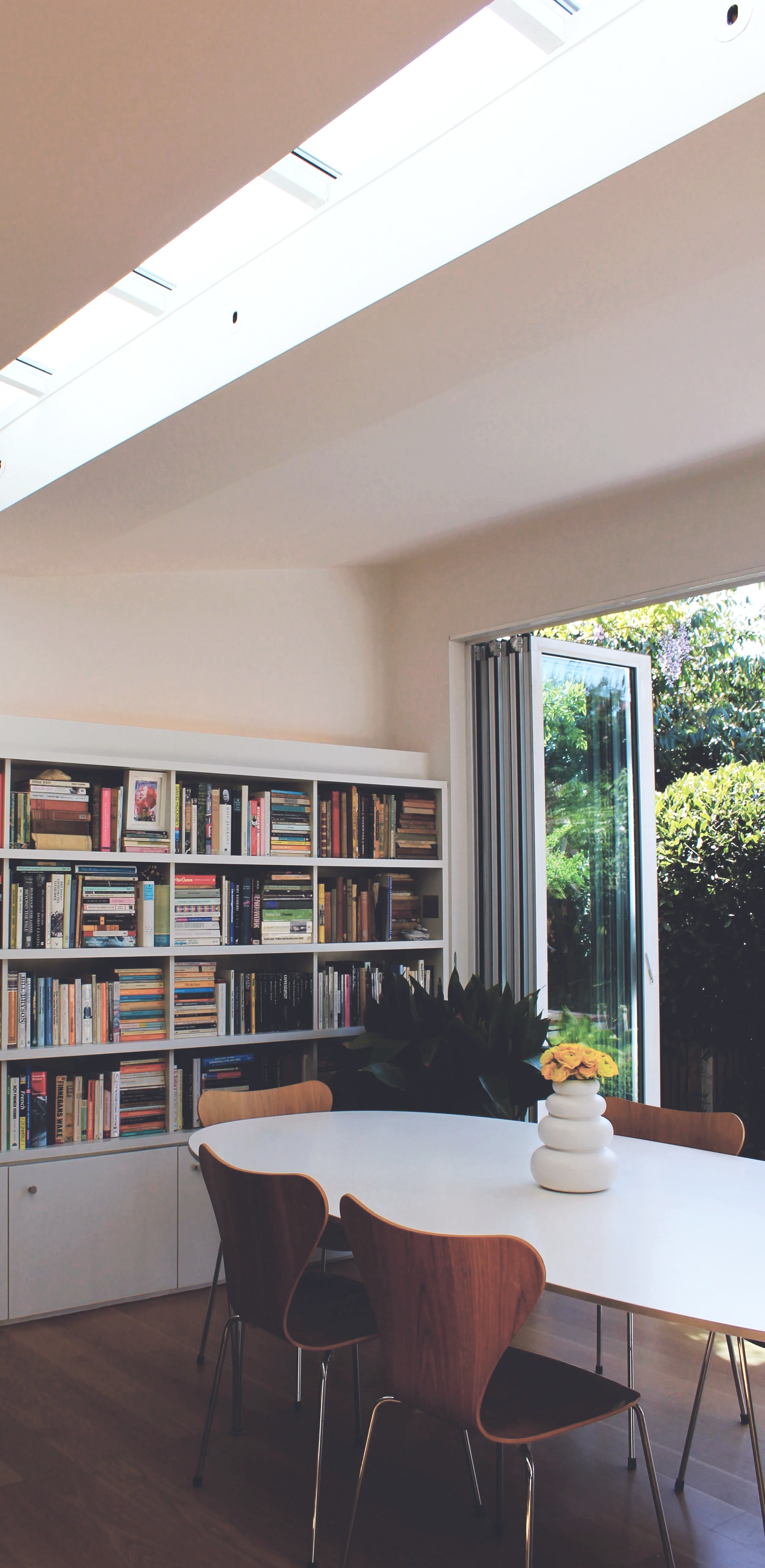 A well-lit room with a white bookshelf filled with books, a white table with a white vase holding yellow flowers, and wooden chairs. A large window with an open glass door reveals greenery outside, and sunlight streams into the room.